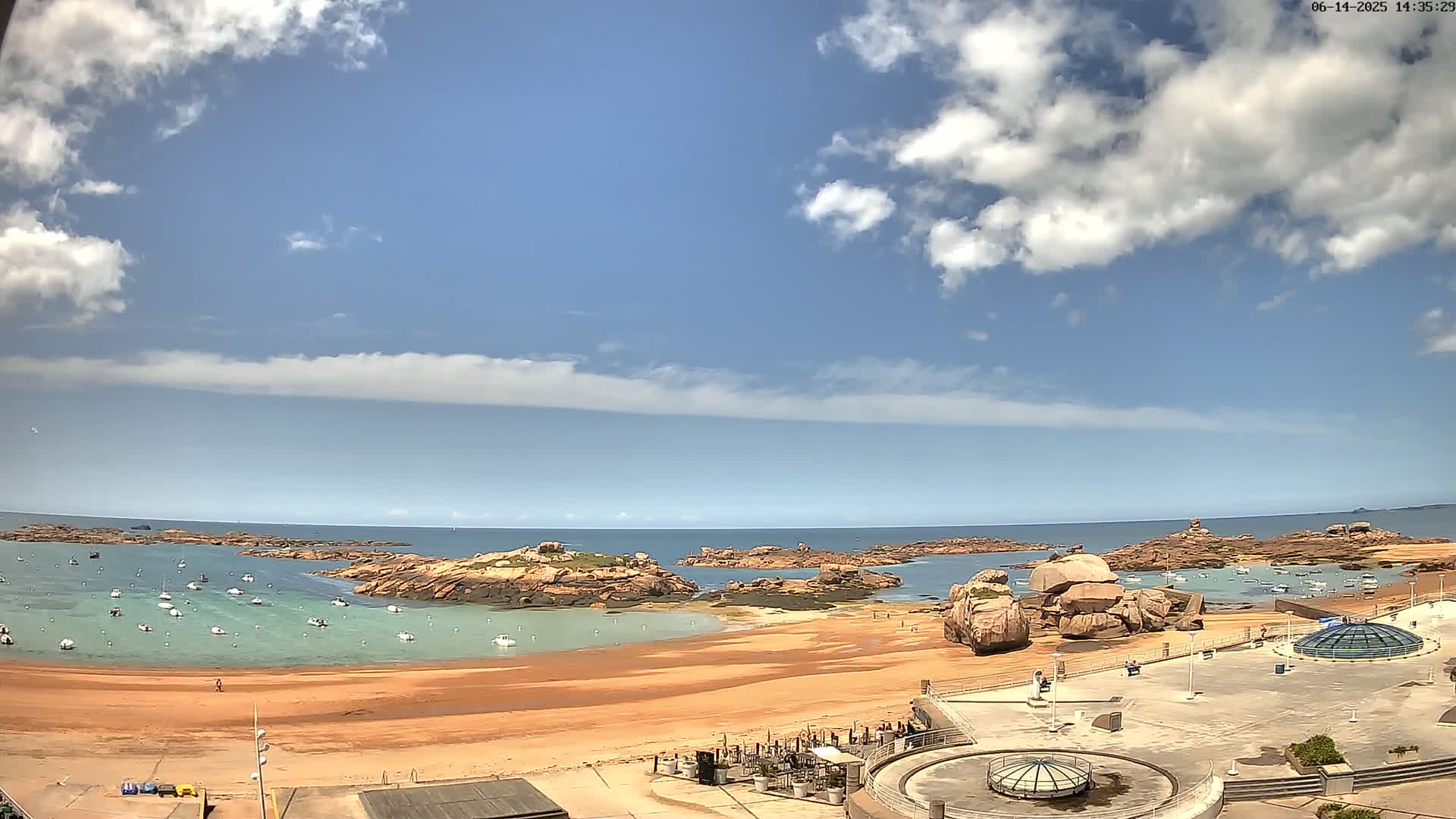 A sandy beach with calm, light-green water, numerous small boats, and pink rocks under a mostly sunny sky with scattered white clouds.