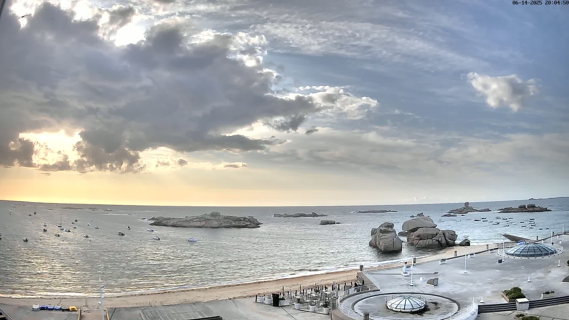 A partly cloudy sunset over a calm ocean with numerous boats and large rocks near a sandy beach and a paved area with structures.