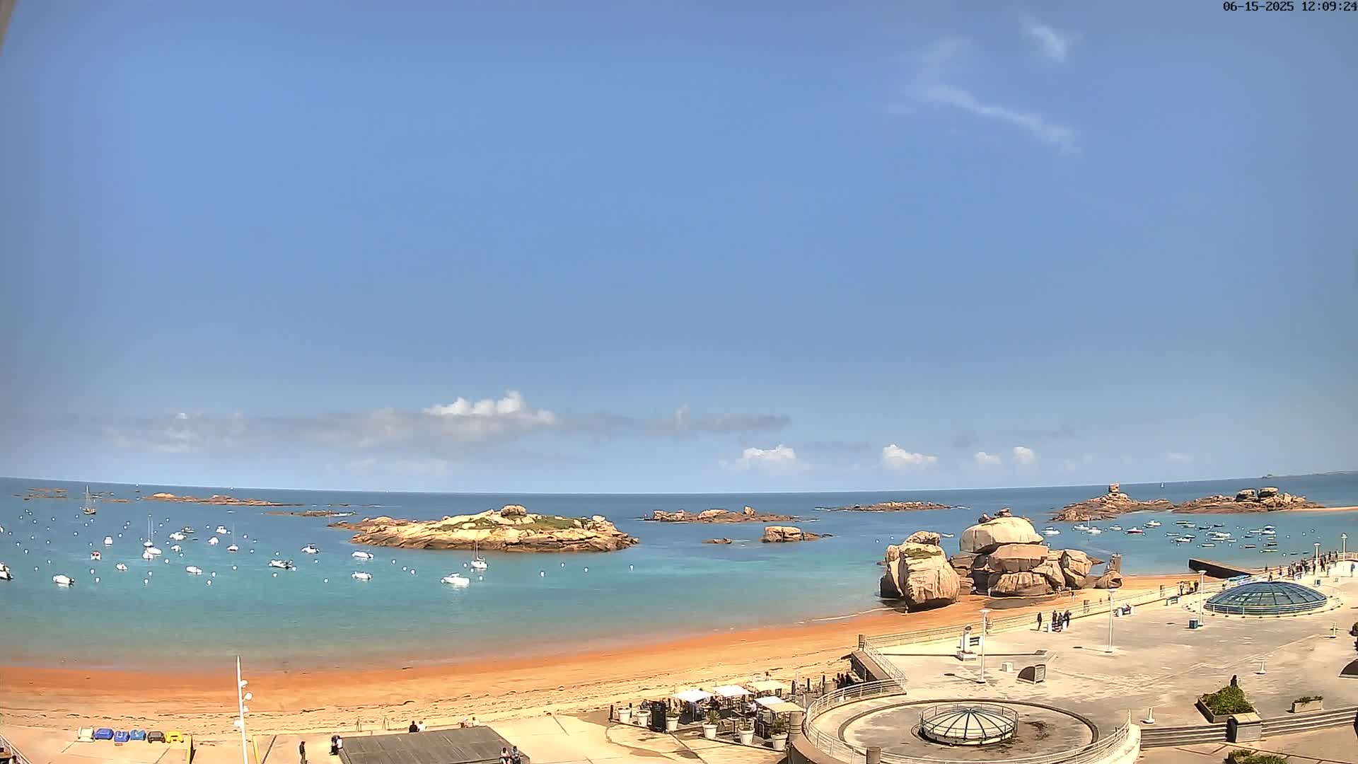 A sandy beach with calm, turquoise water, numerous boats, and rocky outcrops under a mostly sunny sky with a few scattered clouds.