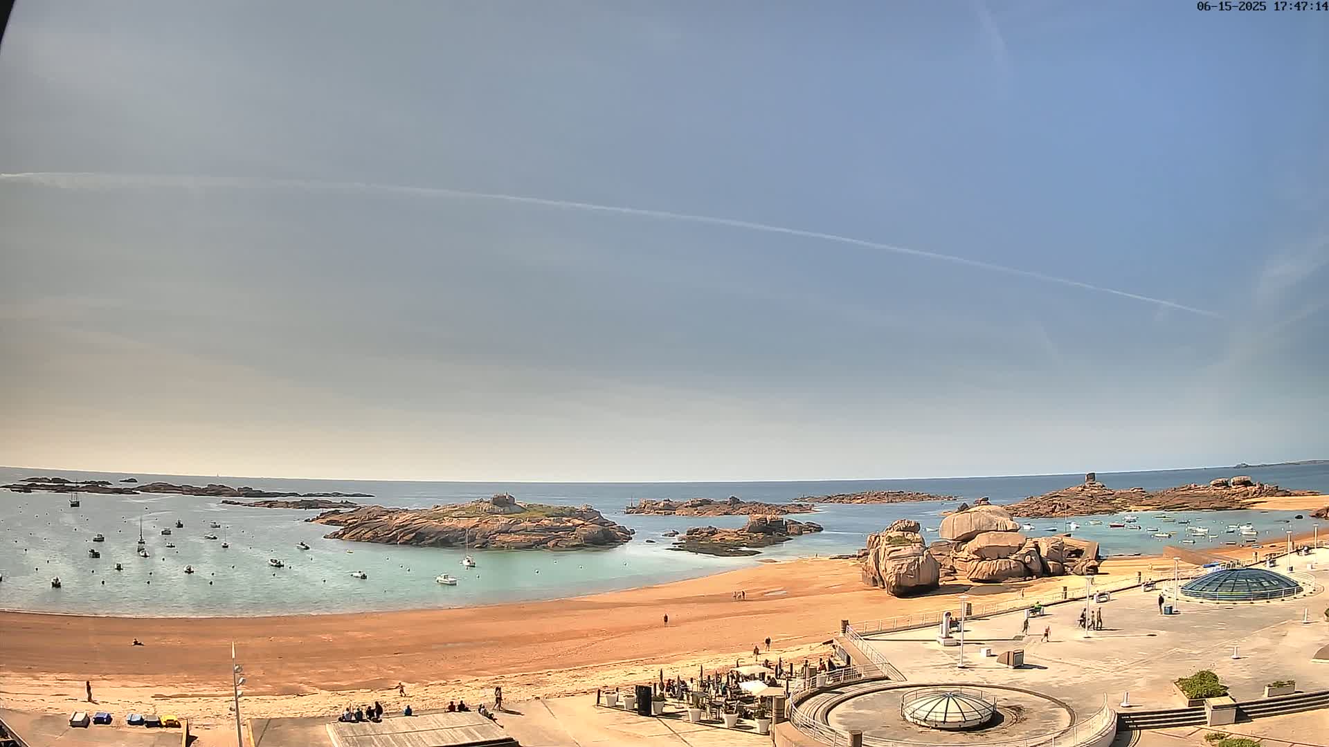 A sandy beach with calm, clear water, numerous small boats, and large pink rocks is visible under a mostly clear, sunny sky.