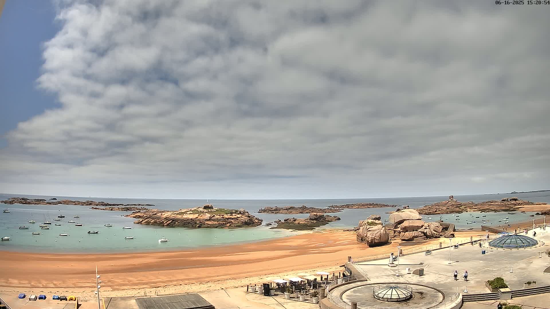 A partly cloudy day reveals a sandy beach with numerous boats in the calm water, surrounded by rocky outcrops and a paved plaza with several people.