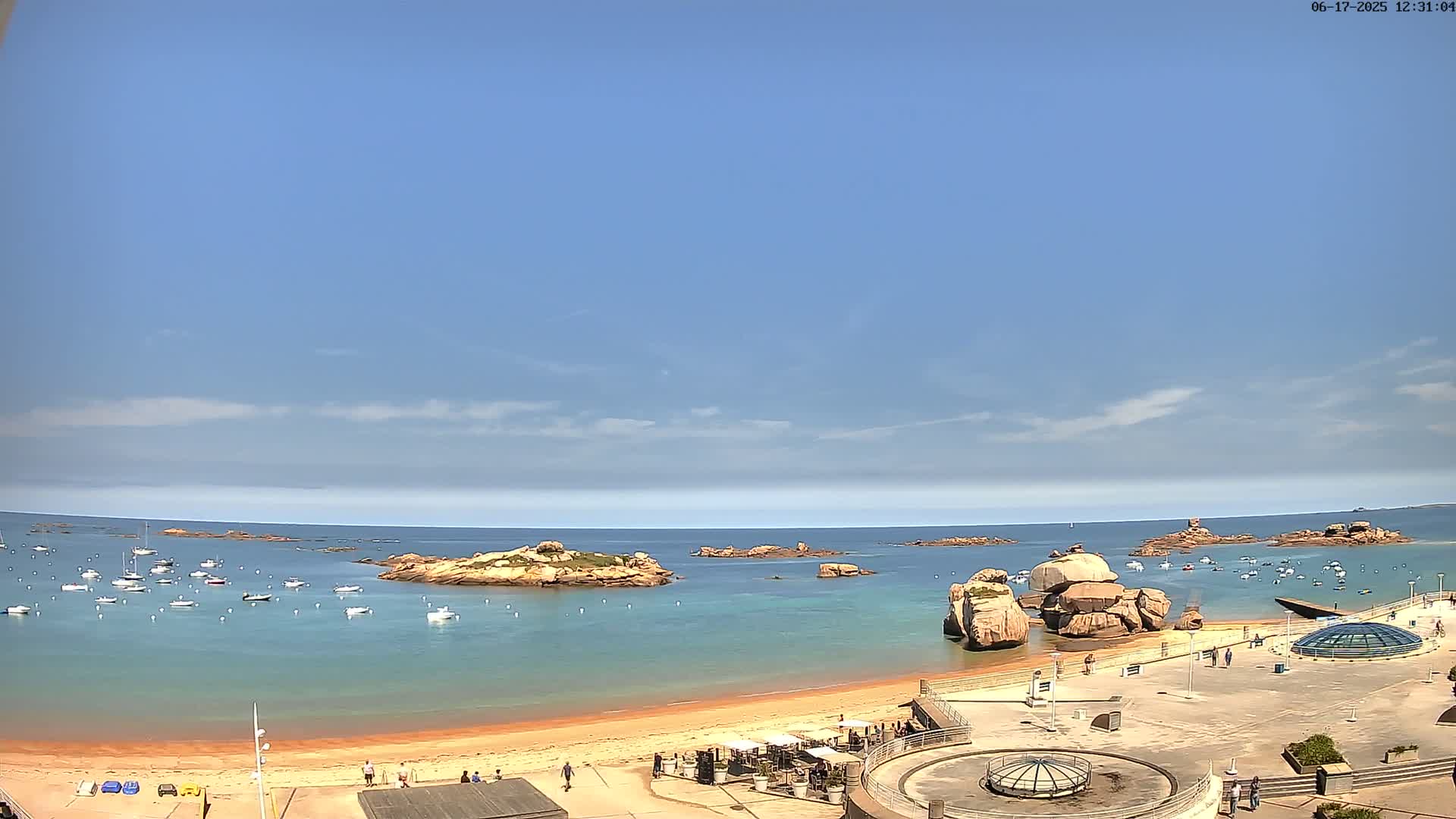 A sandy beach with several boats in the calm, blue water, under a mostly clear sky, is surrounded by rocky outcrops.
