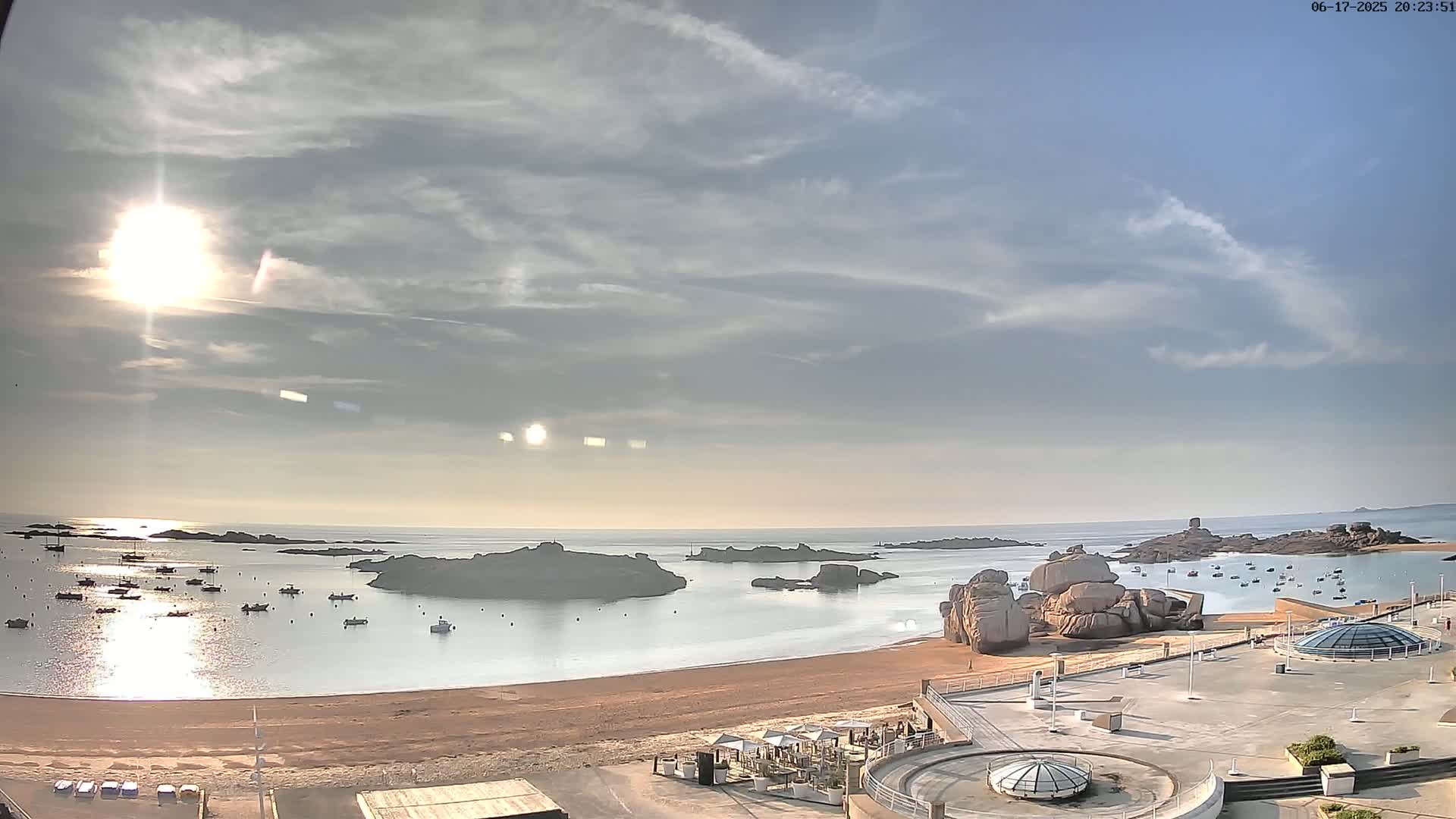 A sandy beach with calm water and numerous boats is visible under a partly cloudy sky, with the sun shining brightly.