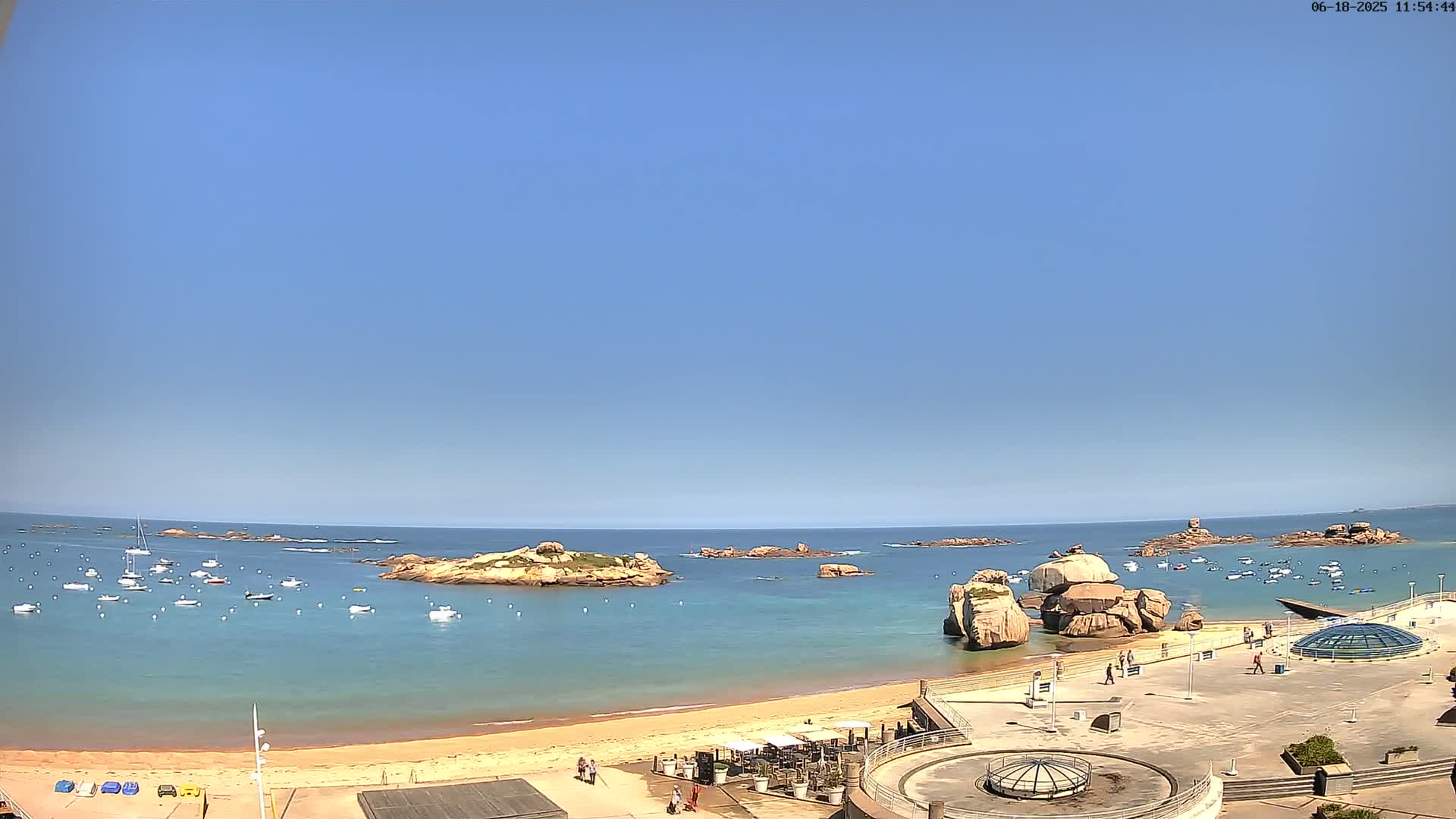 A sandy beach with calm, clear turquoise water, several small rocky islands, and many boats is visible under a clear blue sky.