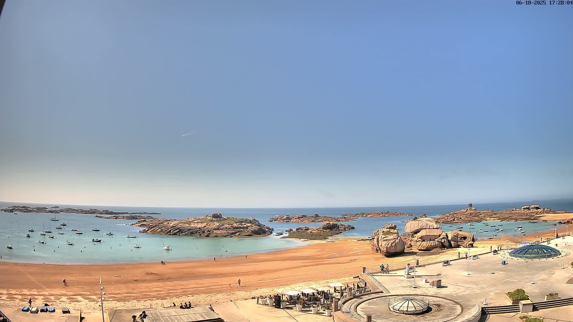 A sandy beach with calm, clear water and several boats is surrounded by rocky outcrops under a clear blue sky.