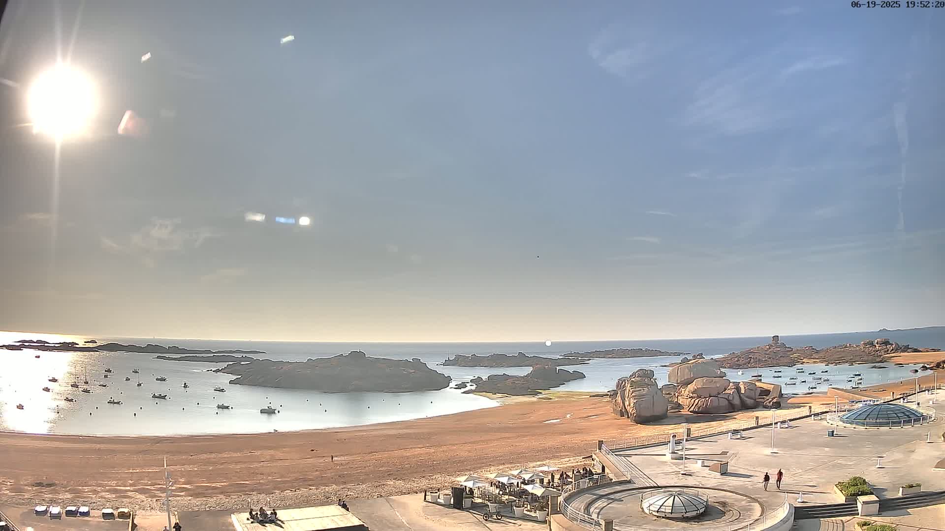 A sunny, mostly clear day overlooks a sandy beach with calm water, numerous small boats, and large rock formations in the distance.
