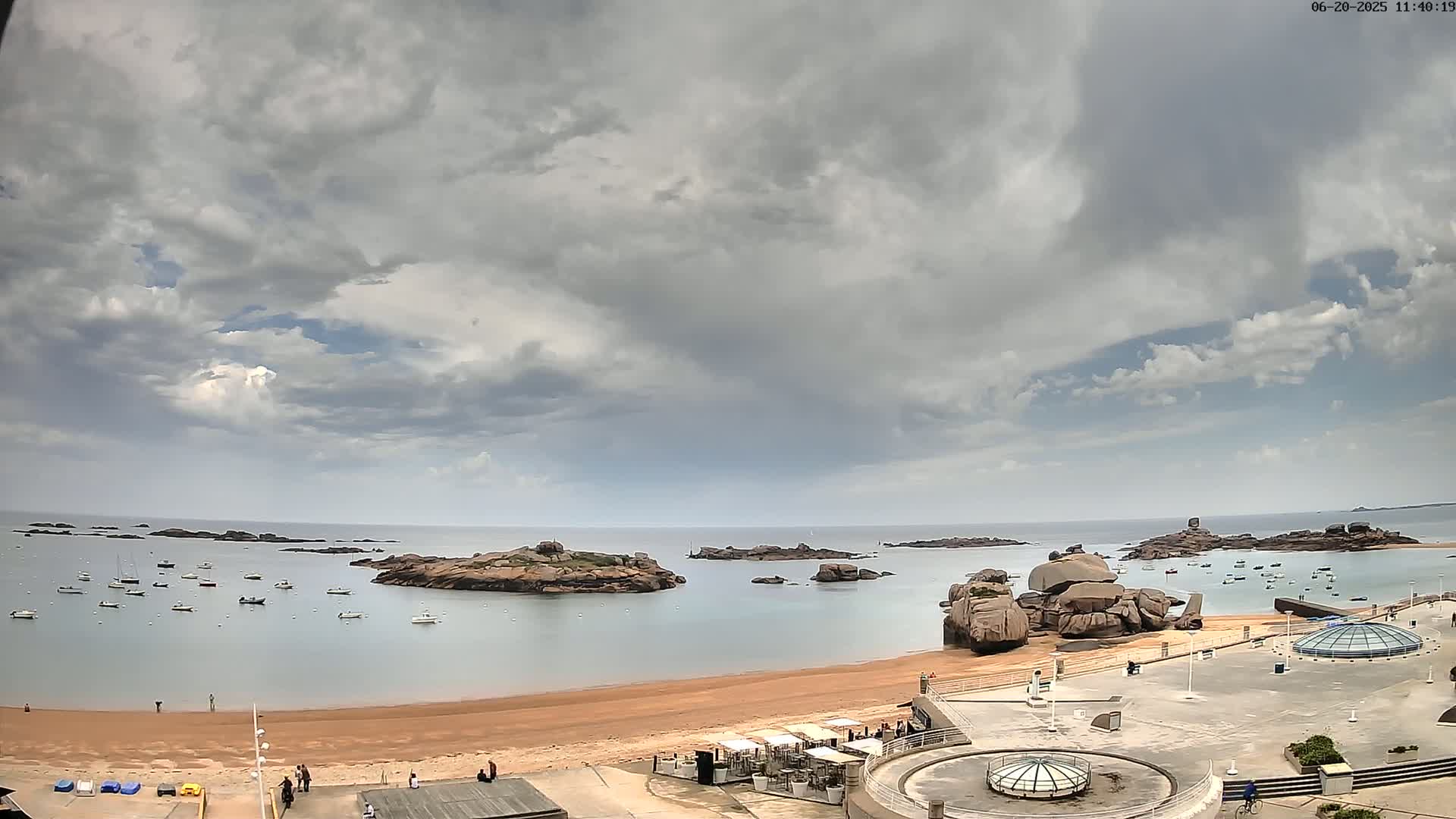 A sandy beach with numerous small boats in the calm water, surrounded by rocky outcrops under a mostly cloudy sky.