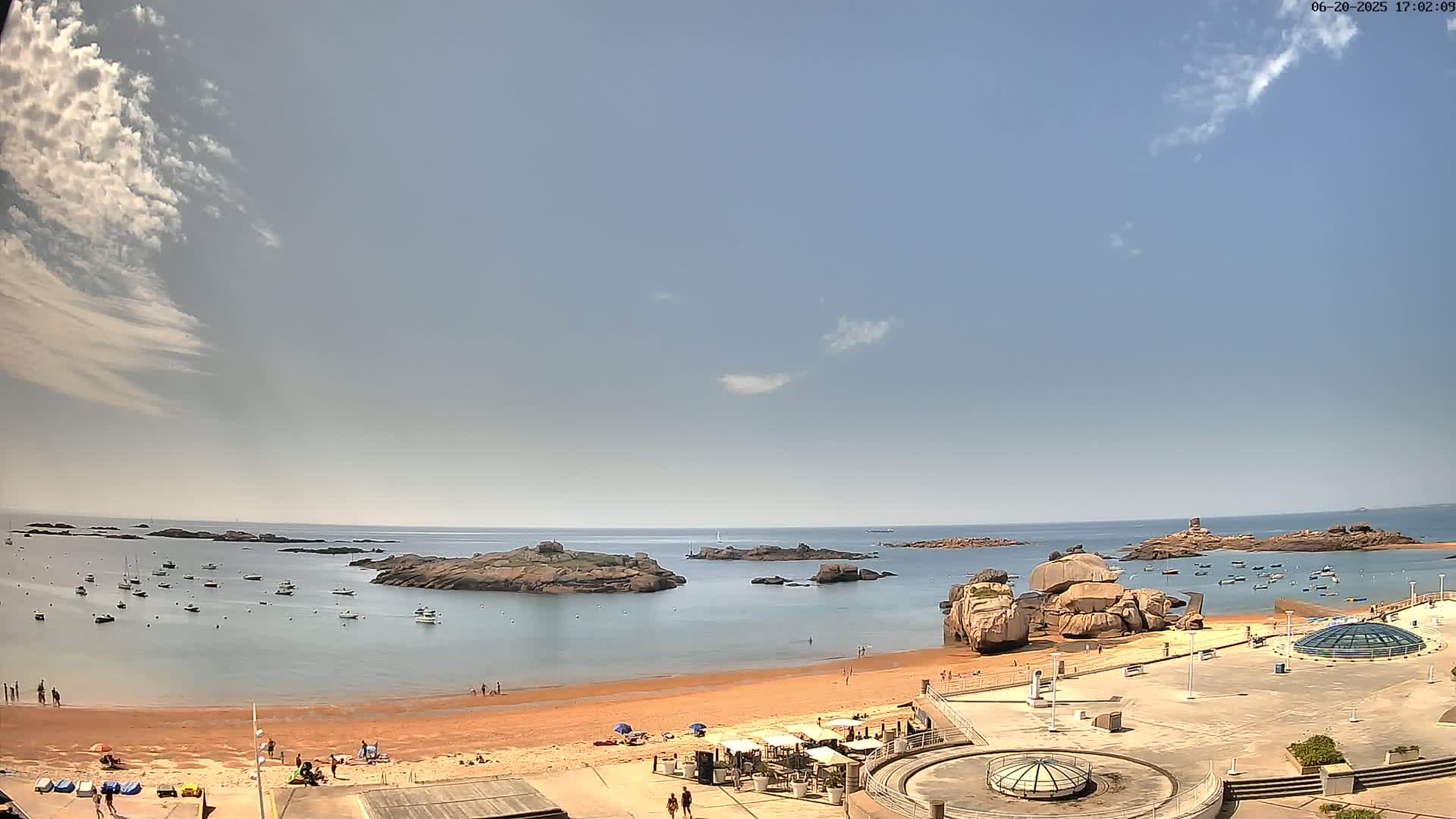 A sandy beach with scattered people and several boats in the calm water, surrounded by rocky islands under a mostly clear sky with a few scattered clouds.