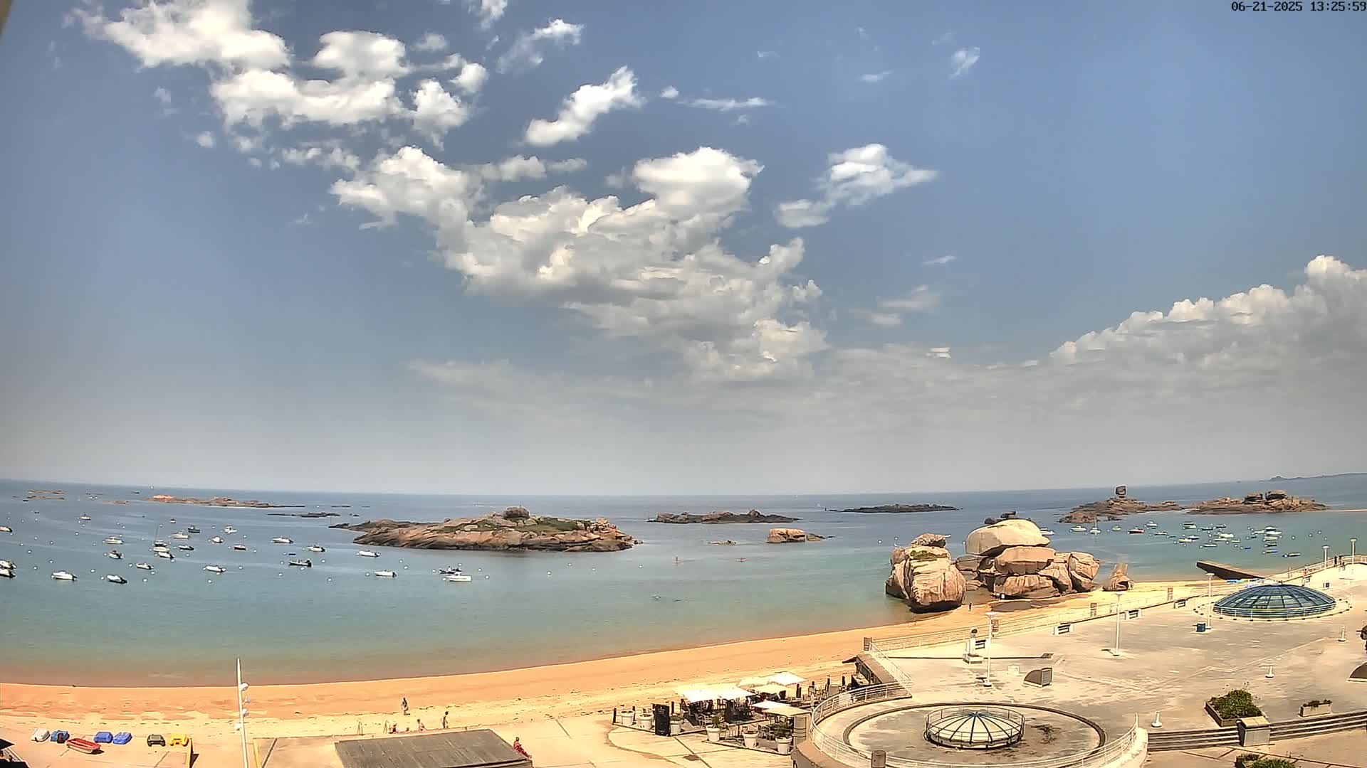 A sandy beach with a calm bay dotted with small boats under a partly cloudy sky.