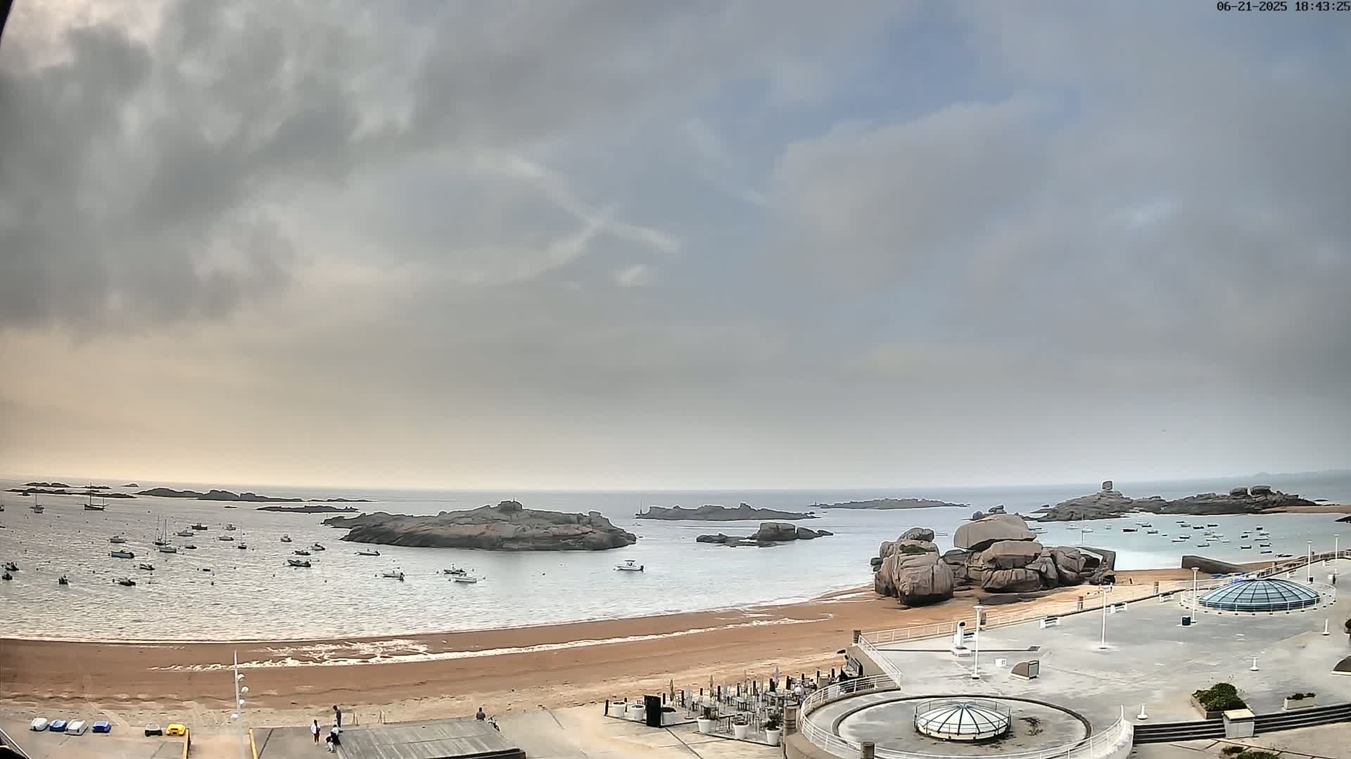 A sandy beach with several boats in the water, rocky outcroppings, and an overcast sky.