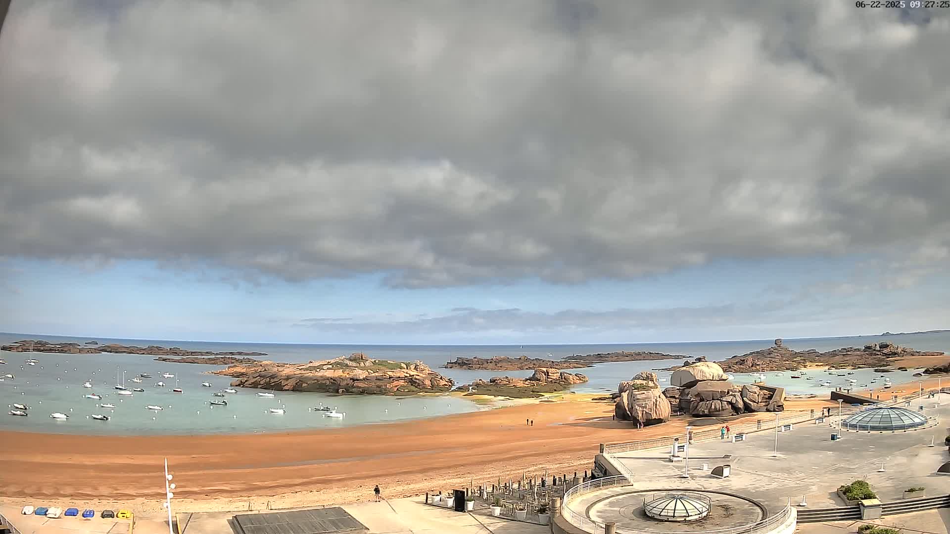 A sandy beach with numerous boats in the calm water, rocky outcrops, and a partly cloudy sky is visible from an elevated vantage point.