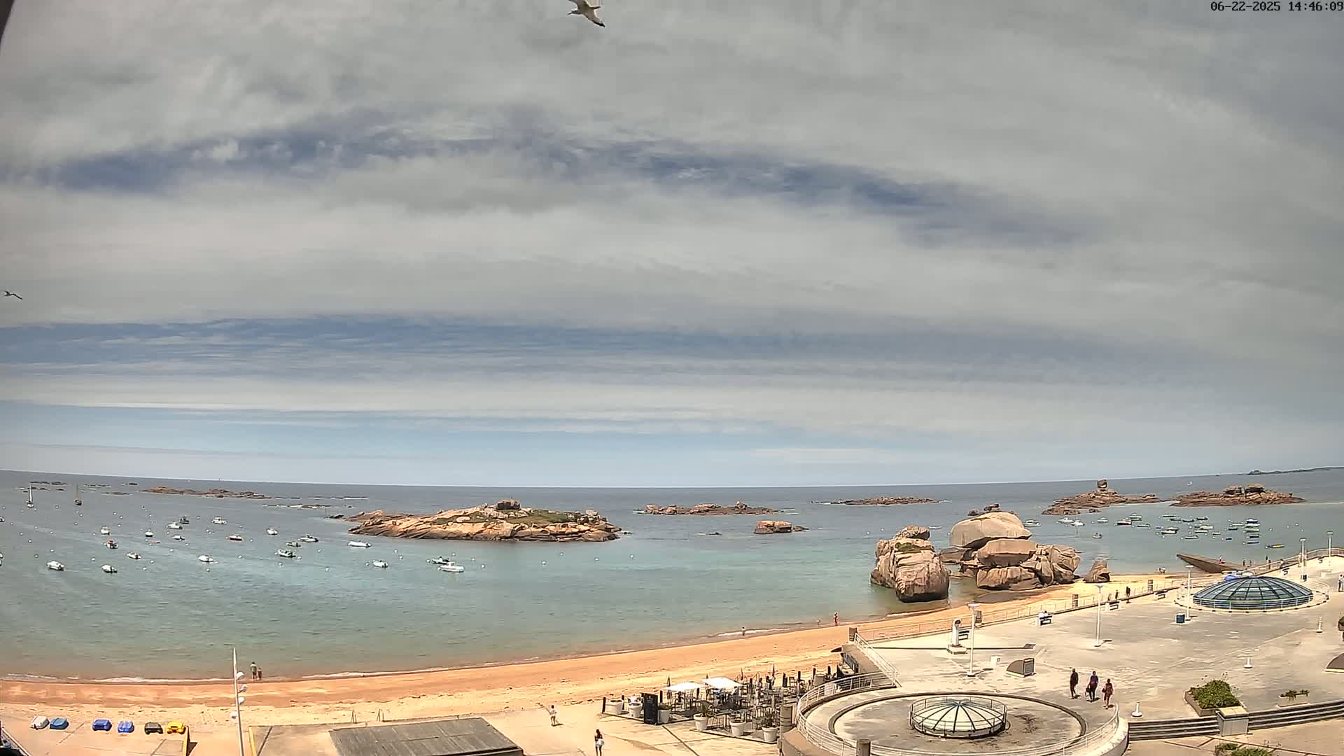 A sandy beach with numerous small boats in the calm water, surrounded by rocky outcrops under a mostly cloudy sky.