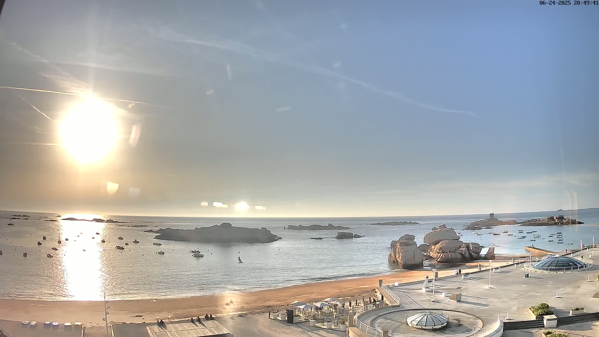 A bright, sunny day at a coastal location shows a sandy beach, calm water dotted with boats, rocky outcrops, and a paved area with structures.