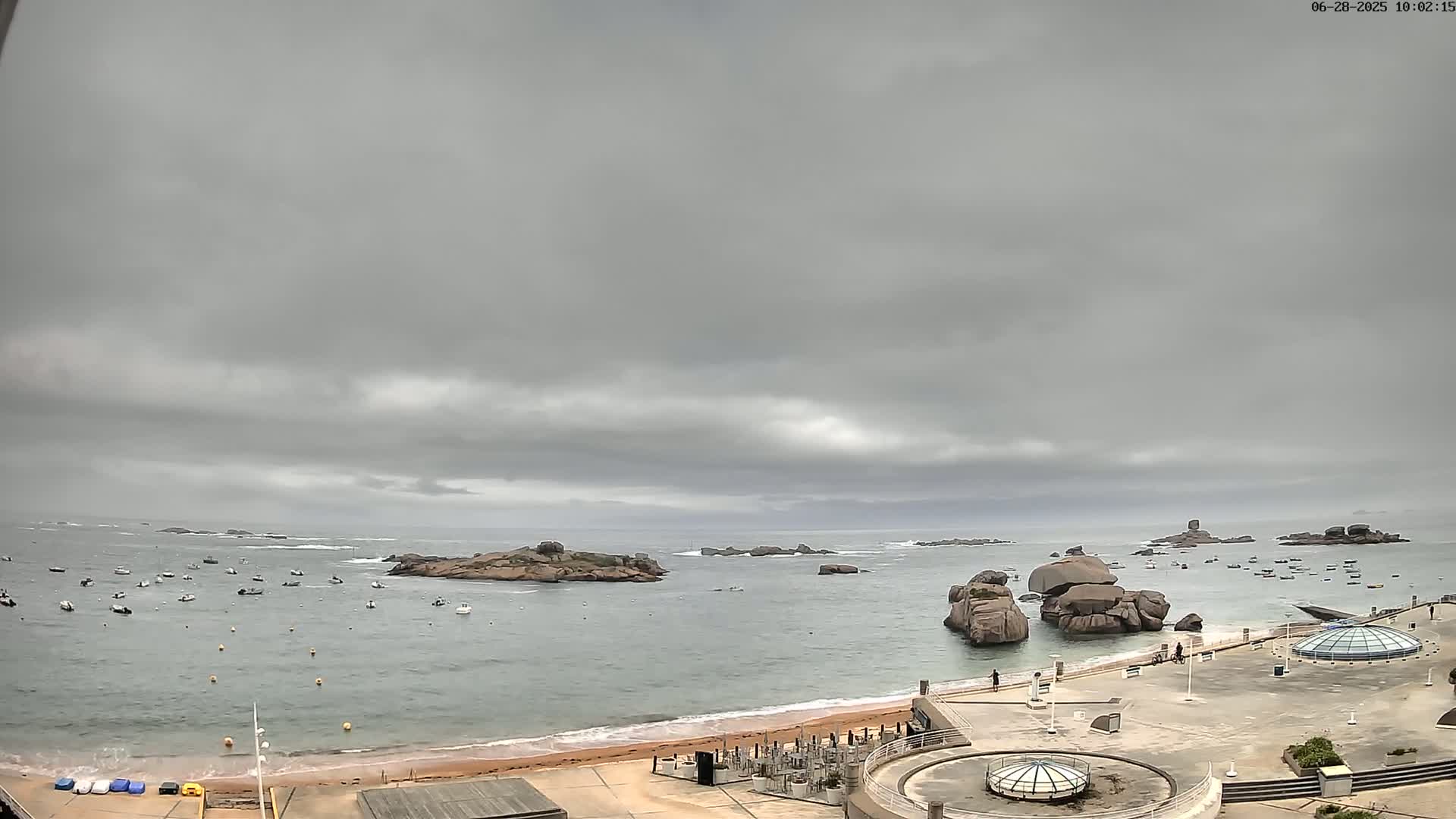 A mostly cloudy day overlooks a calm bay with numerous small boats, rocky outcroppings, a sandy beach, and a paved area with structures.