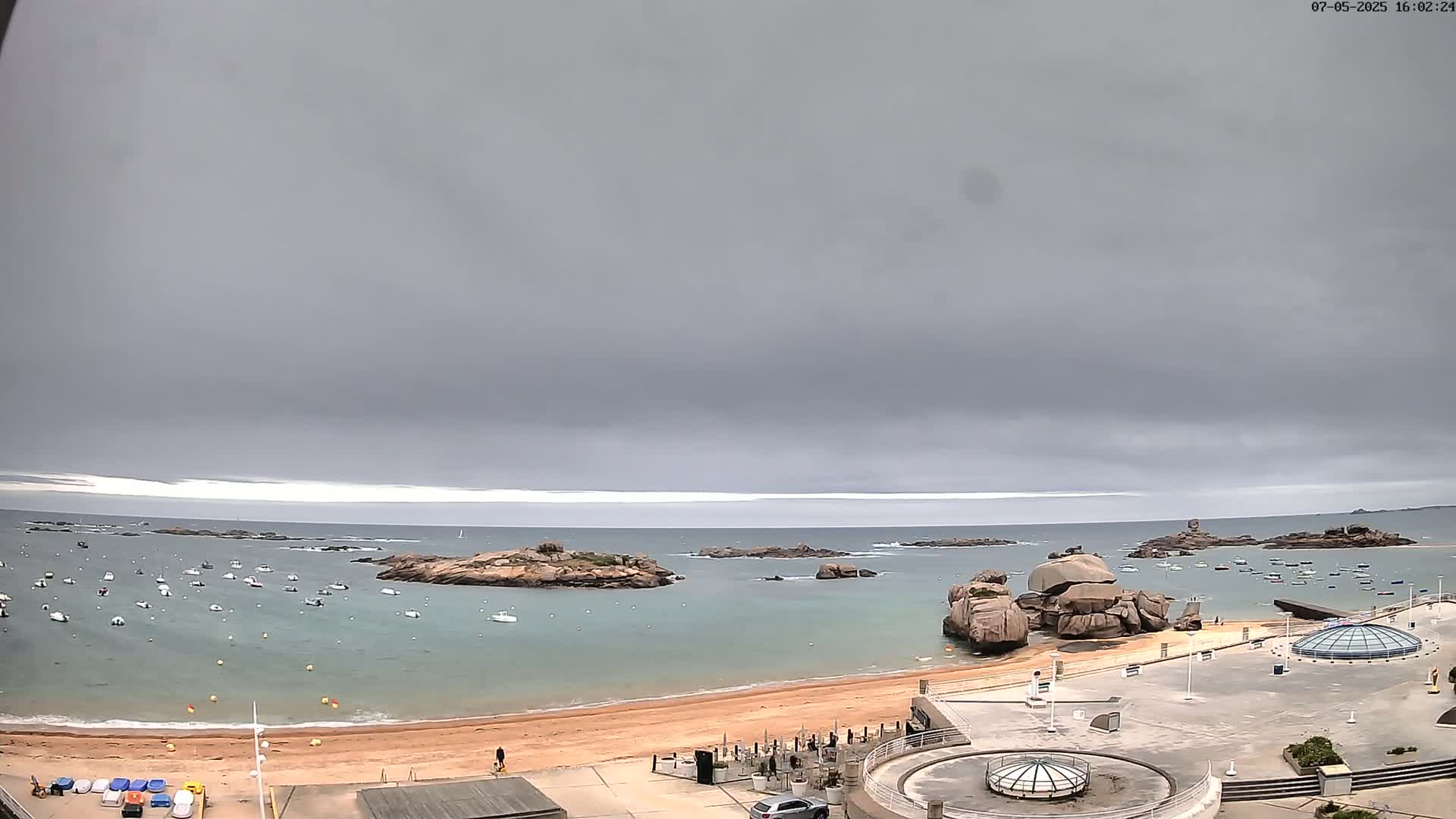 A sandy beach with several boats in the calm water is surrounded by rocky outcrops under an overcast sky.