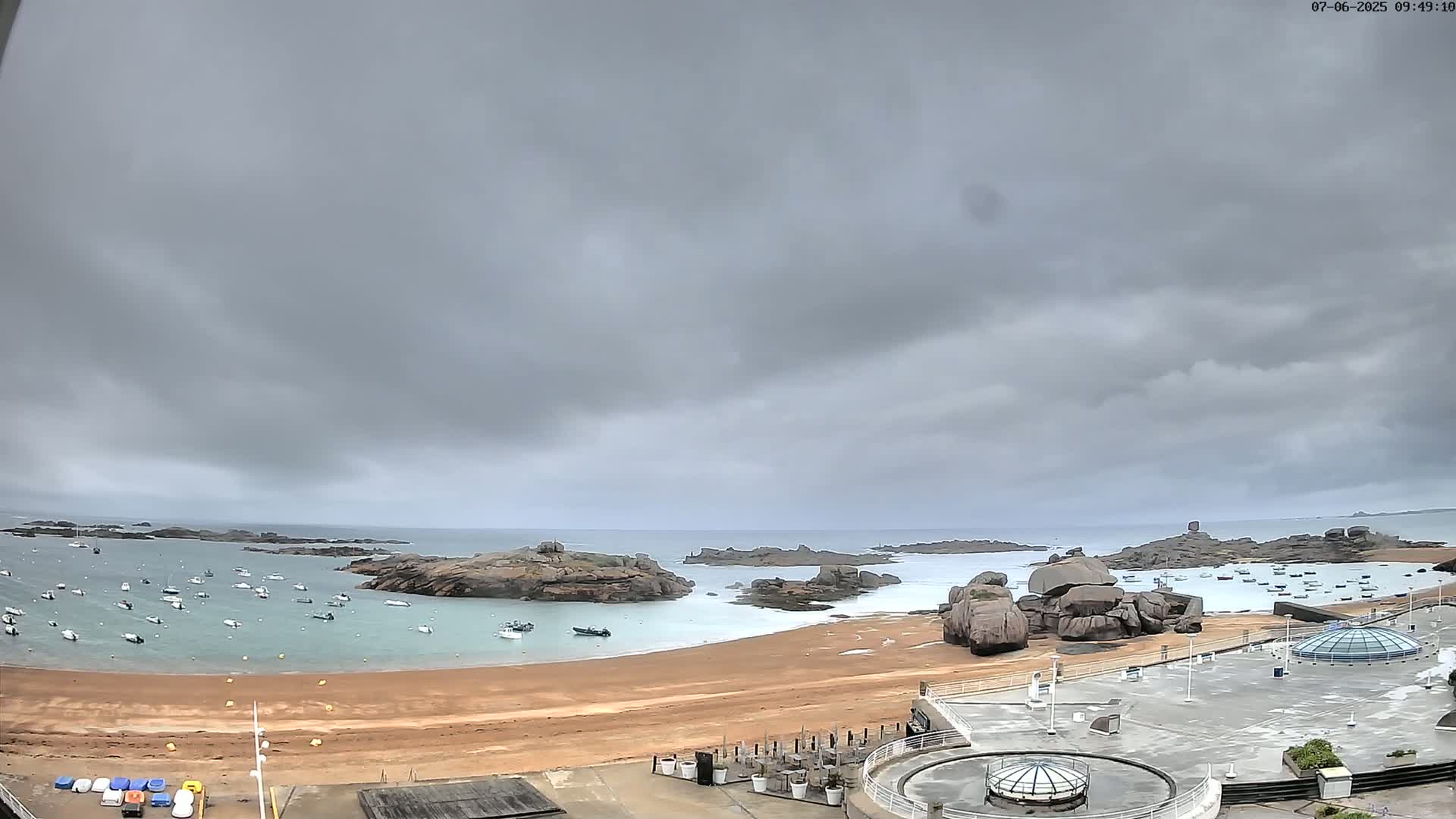 A sandy beach with numerous small boats in calm water, backed by rocky outcroppings under an overcast sky.