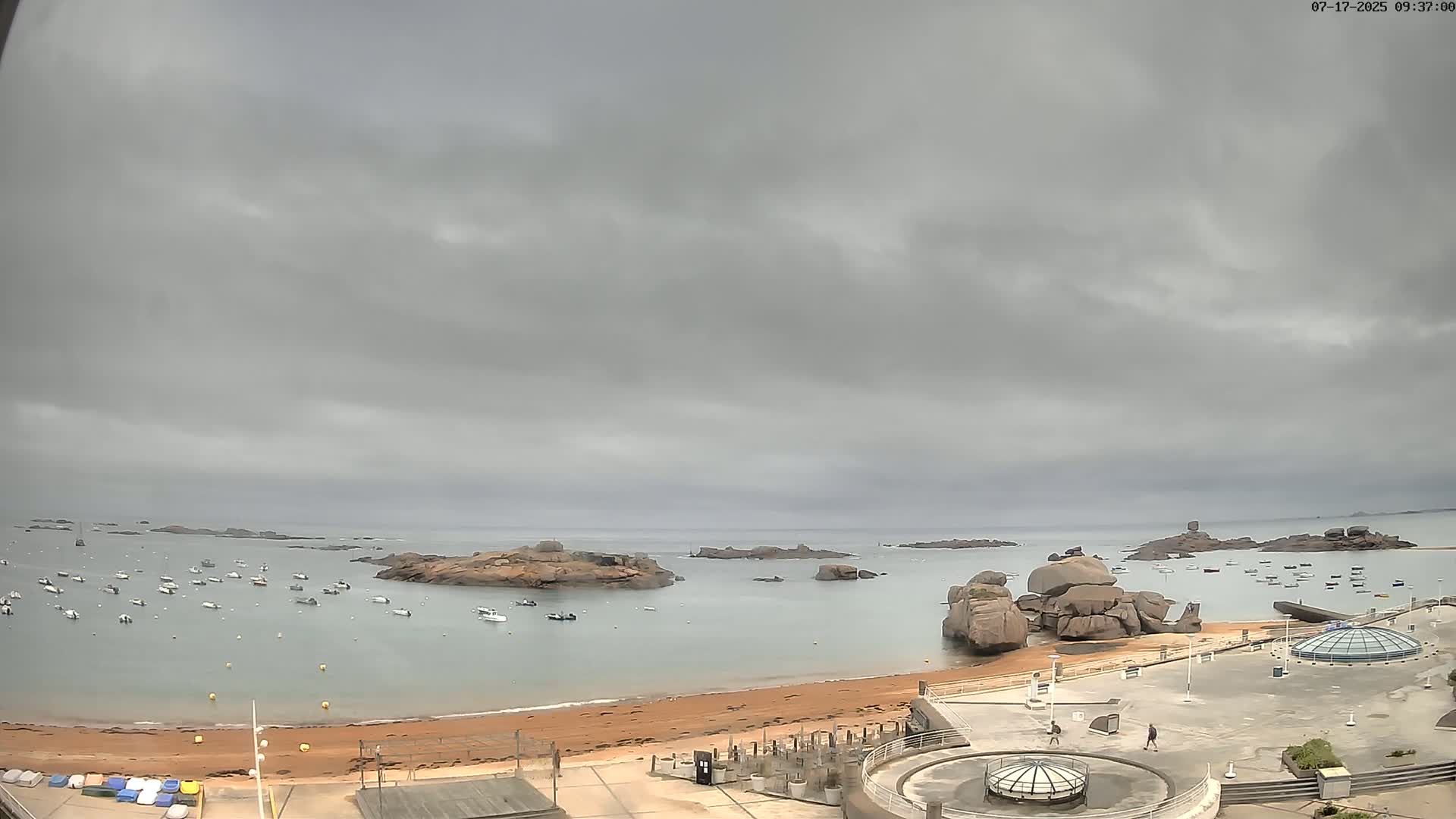 A calm bay with numerous small boats and large rocks, under an overcast sky, is viewed from an elevated position overlooking a sandy beach and paved area.