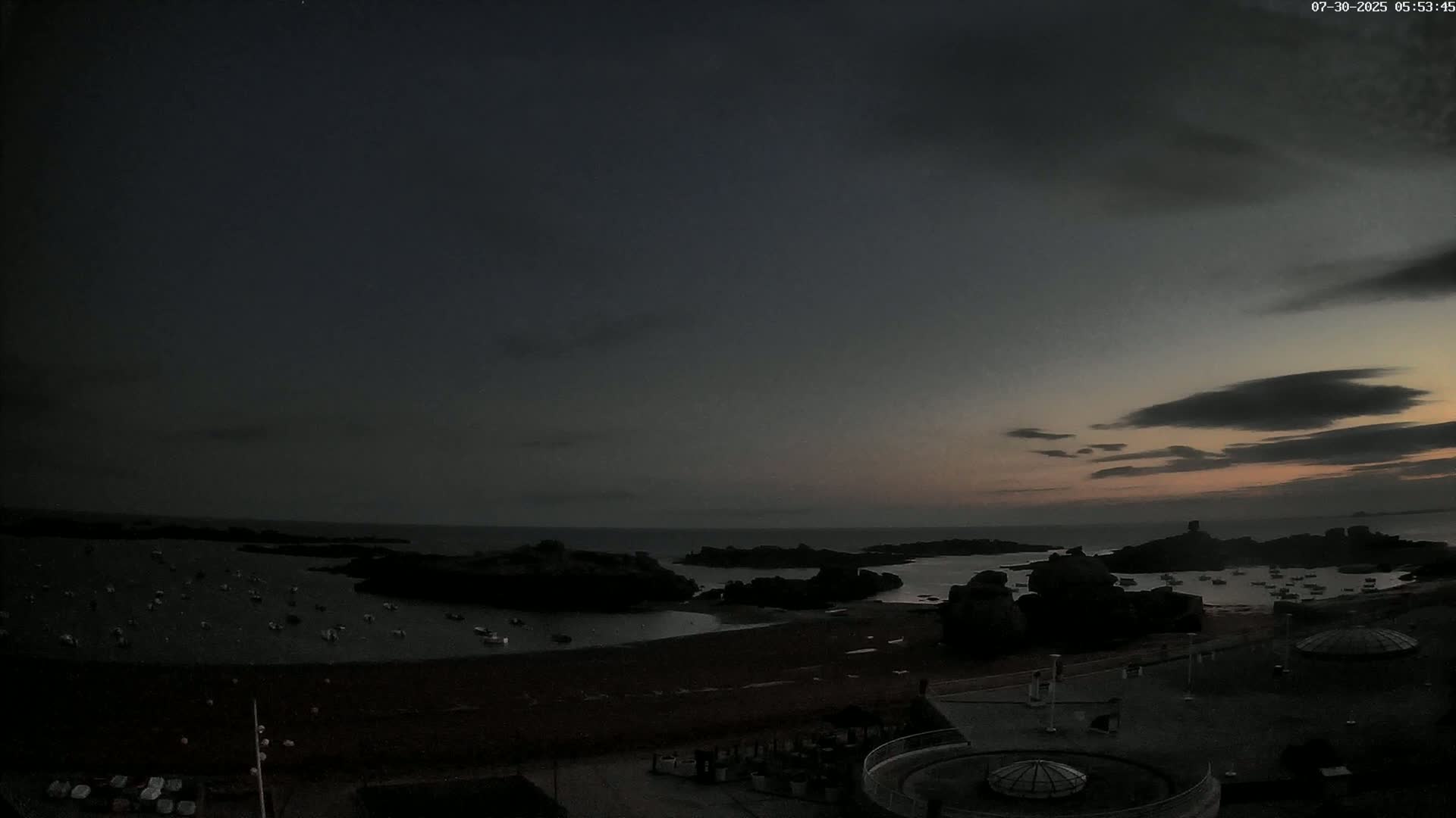 A calm bay with numerous small boats and large rocks, under an overcast sky, is viewed from an elevated position overlooking a sandy beach and paved area.