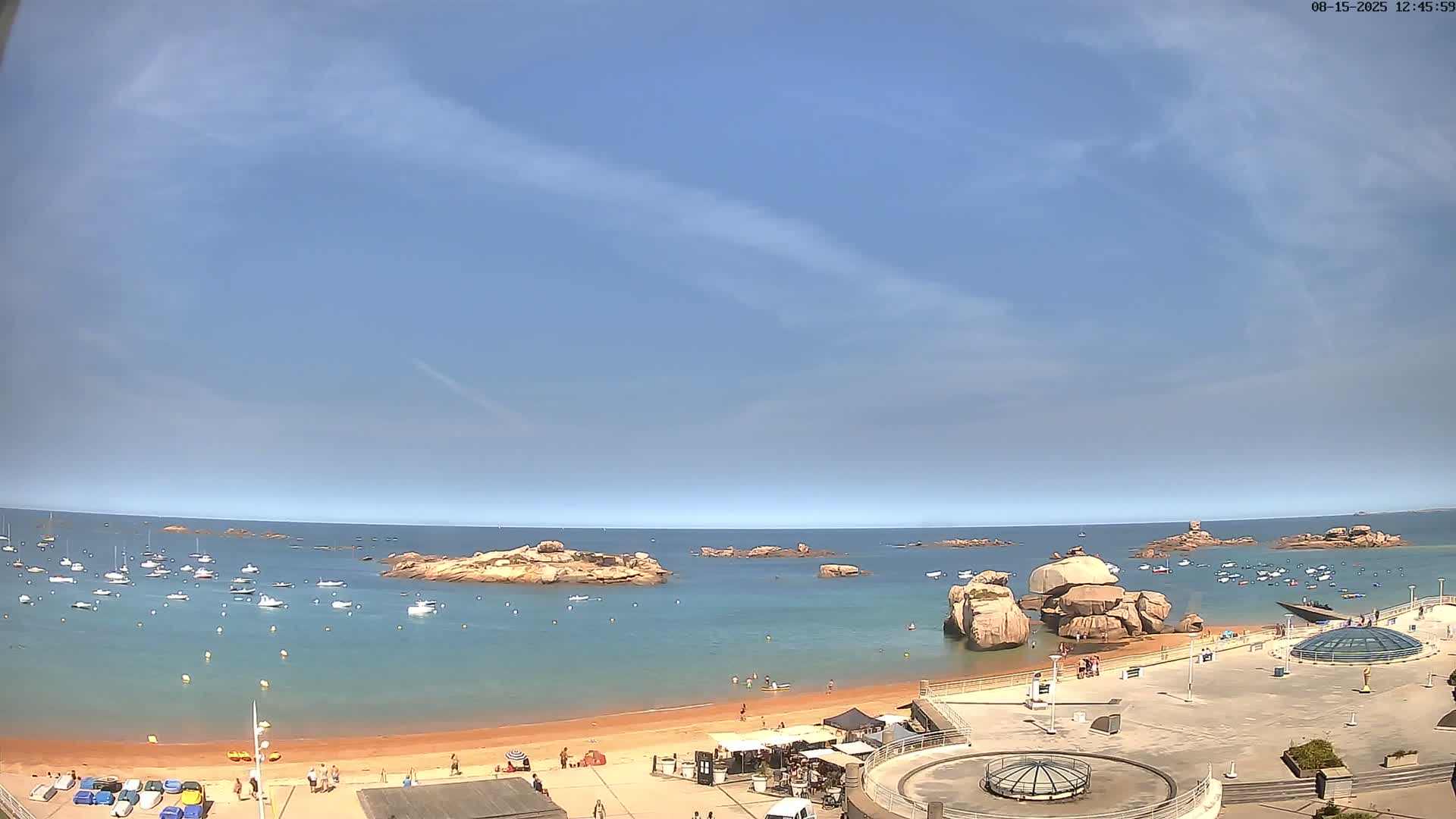A calm bay with numerous small boats and large rocks, under an overcast sky, is viewed from an elevated position overlooking a sandy beach and paved area.