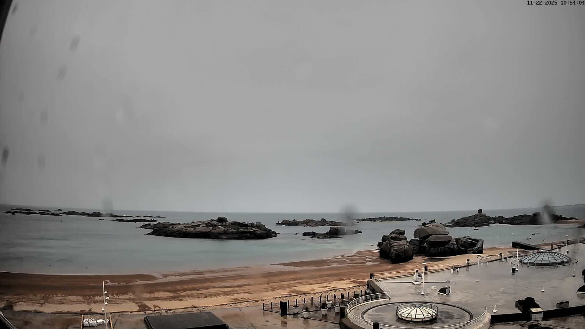 The image depicts an overcast and rainy coastal scene featuring a rocky shoreline, a wet sandy beach, and a modern waterfront promenade with circular glass structures, all under a uniformly gray sky.