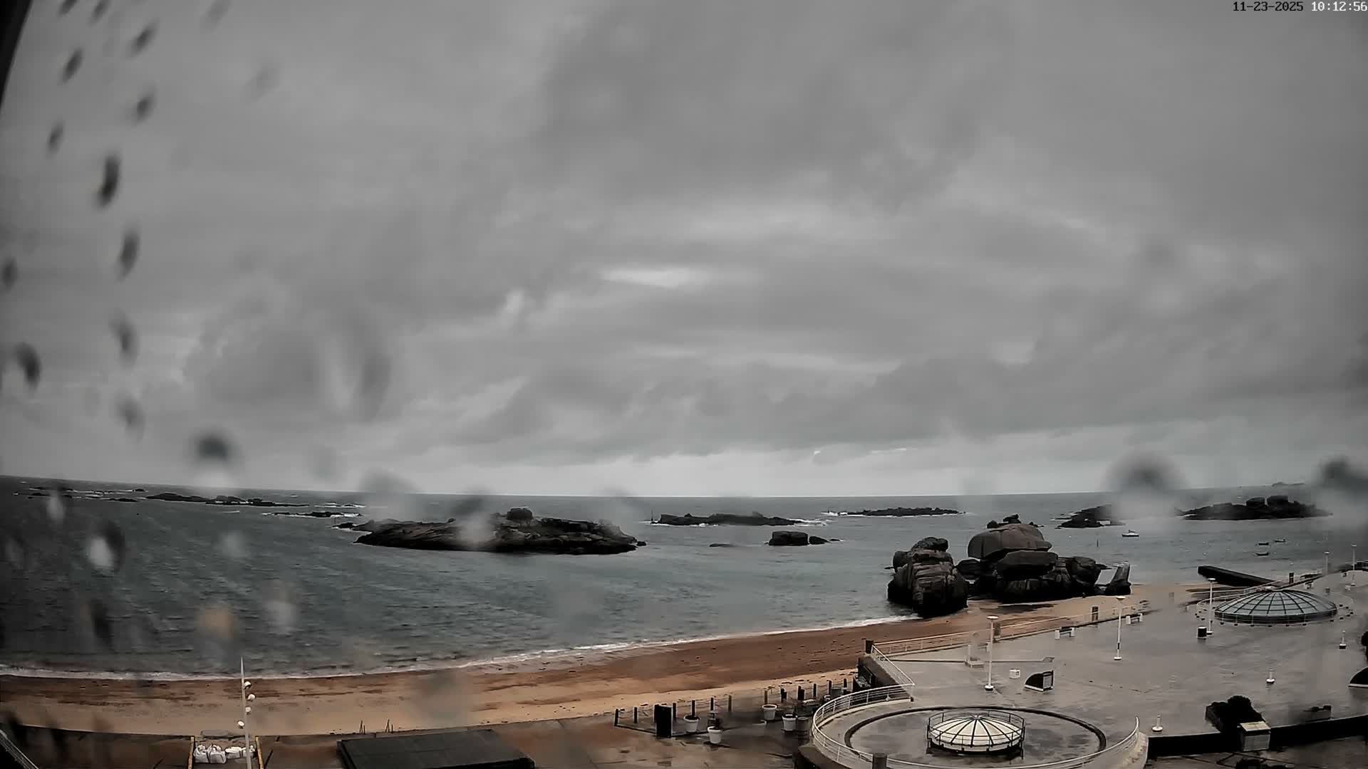 An overcast and rainy coastal scene features a sandy beach, dark choppy sea with numerous rocky islets, and a wet concrete promenade with dome-shaped structures, all viewed through a rain-streaked lens.