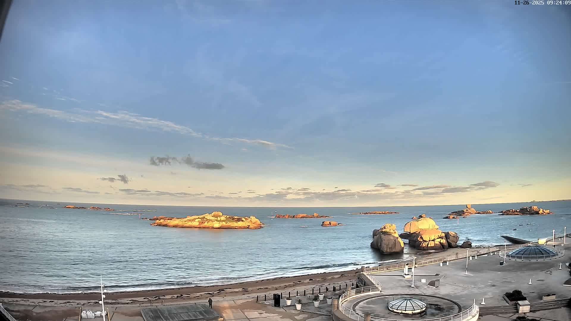 A wide coastal view under a mostly clear blue sky with scattered clouds, showcasing a calm blue sea dotted with numerous orange-tinted rocky islets, a sandy beach, and a modern waterfront promenade.