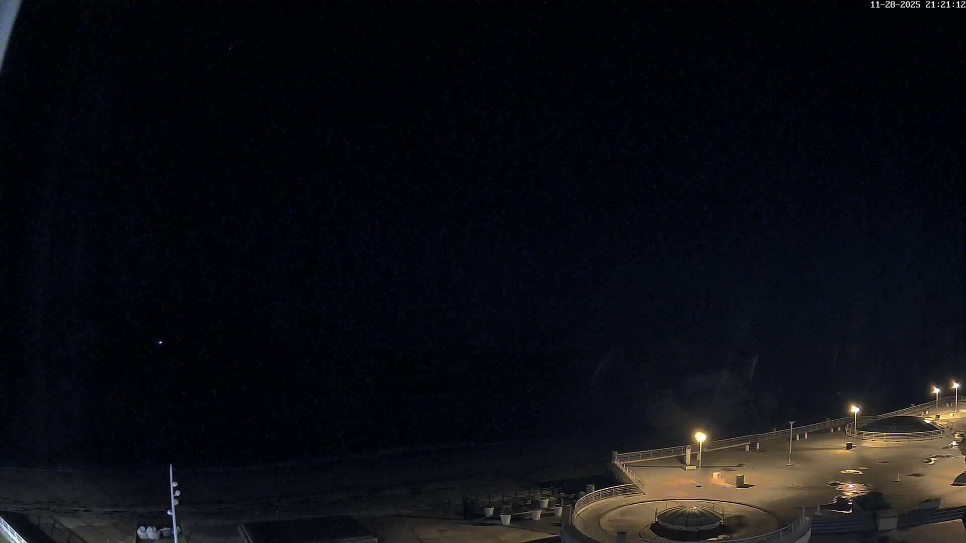 A brightly lit promenade with wet surfaces, indicating recent rain, extends alongside a dark ocean under a deep night sky.