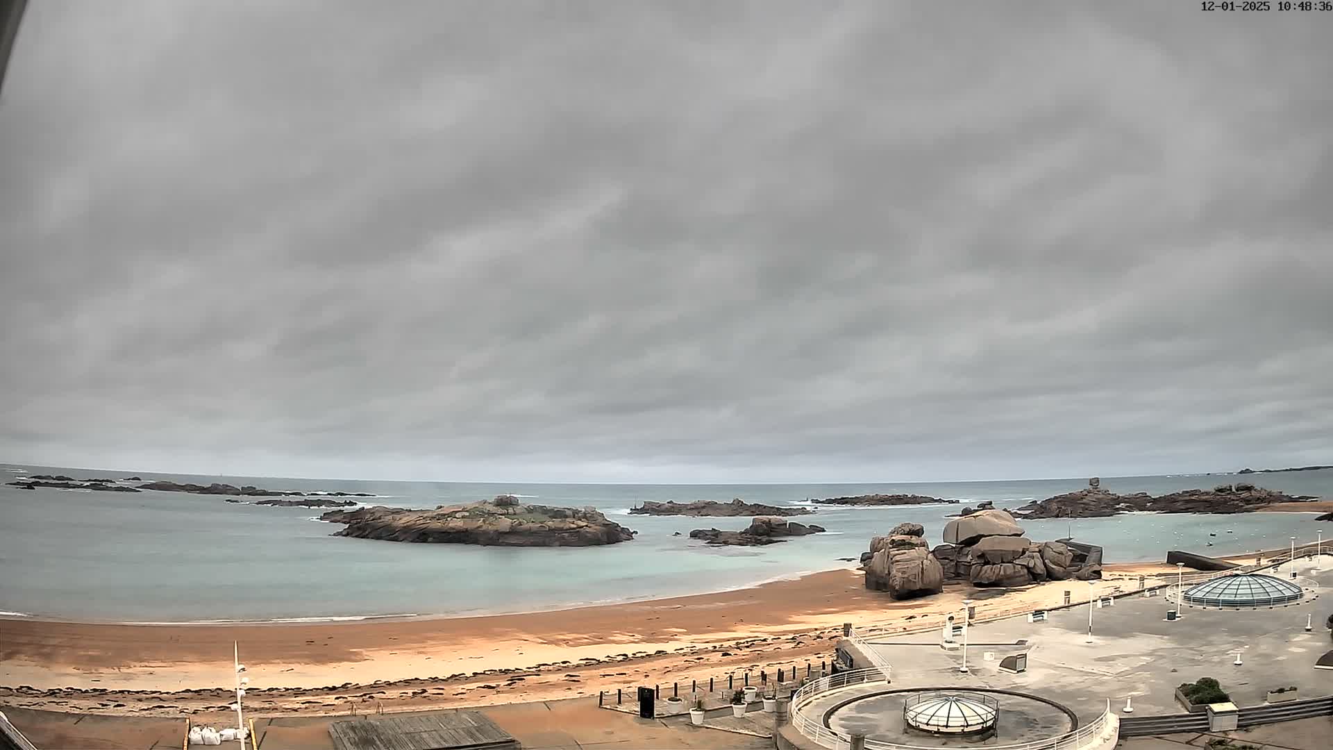 A wide shot captures a coastal scene featuring a sandy beach, distinctive massive rock formations protruding from the calm, light blue-green sea and along the shore, and a paved waterfront area with modern domed structures in the foreground, all under a uniform, grey overcast sky.