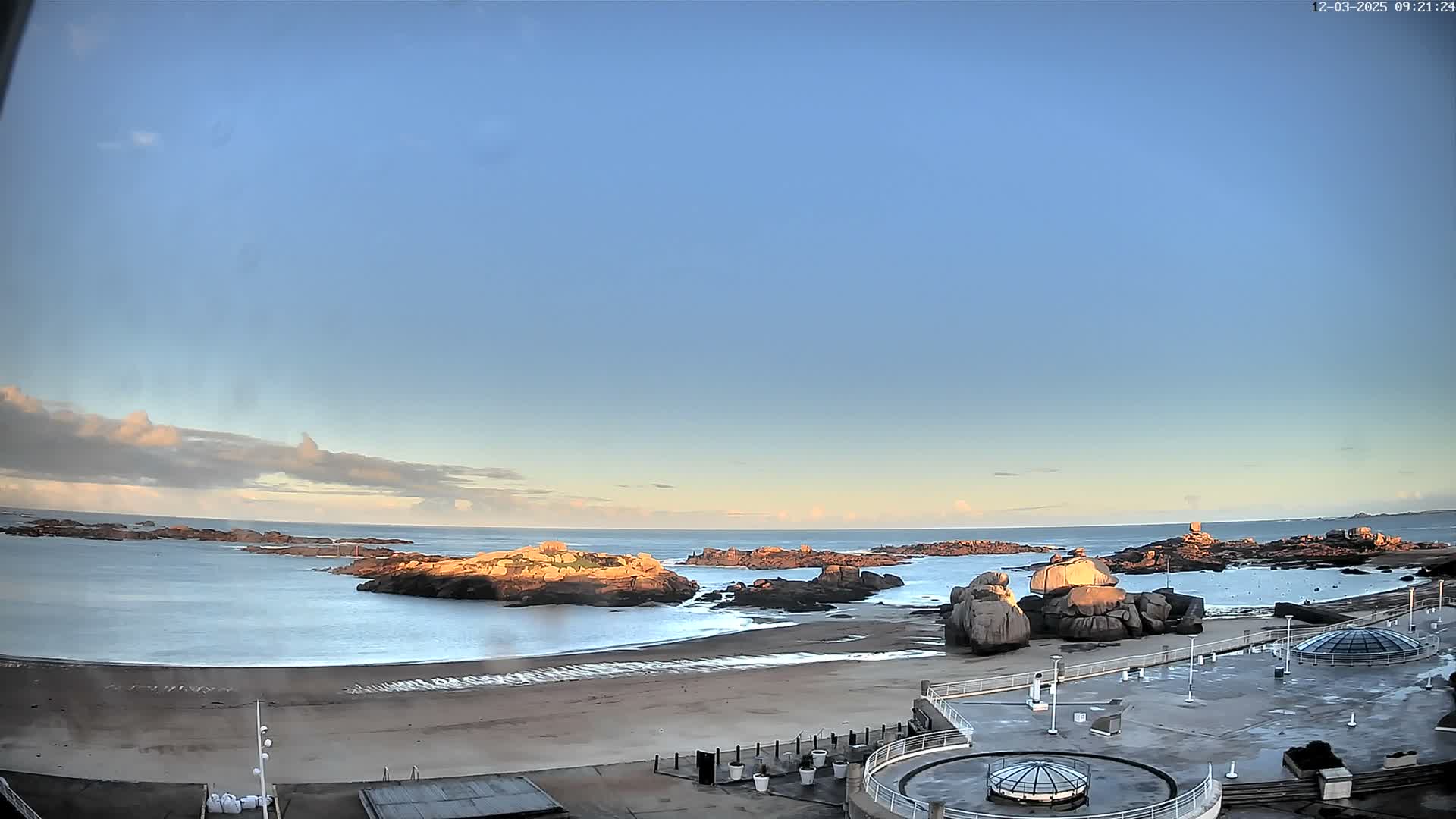 A panoramic view captures a rocky coastline with a calm blue ocean, a wide sandy beach, and numerous large rock formations, while a modern concrete promenade with domed skylights occupies the foreground, all under a partly cloudy sky with soft golden light indicating clear and calm weather.