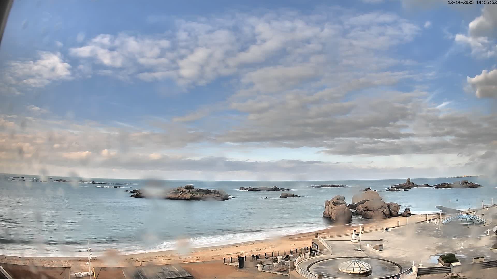 A panoramic view captures a rocky coastline with a calm blue ocean, a wide sandy beach, and numerous large rock formations, while a modern concrete promenade with domed skylights occupies the foreground, all under a partly cloudy sky with soft golden light indicating clear and calm weather.