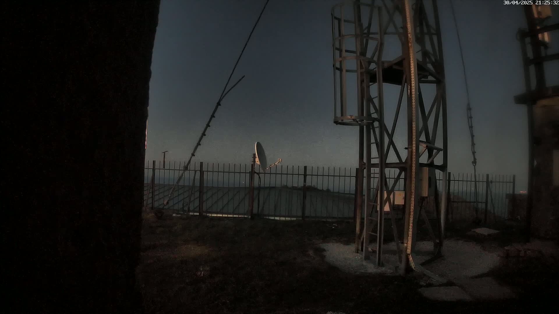 A metal tower with a satellite dish stands behind a fence on a dark, twilight evening.