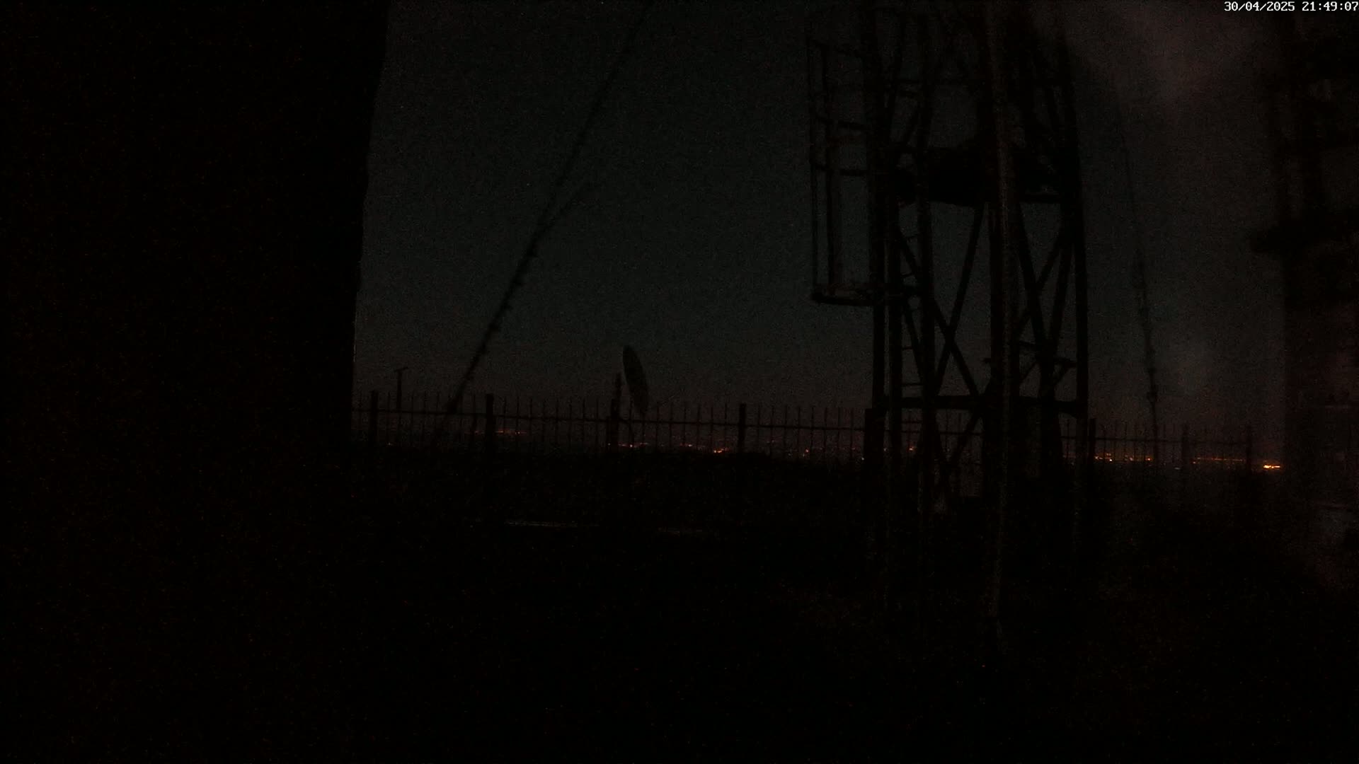 A dark nighttime scene shows a metal structure in the foreground, a fence, and a distant cityscape with faint lights under a dark sky.