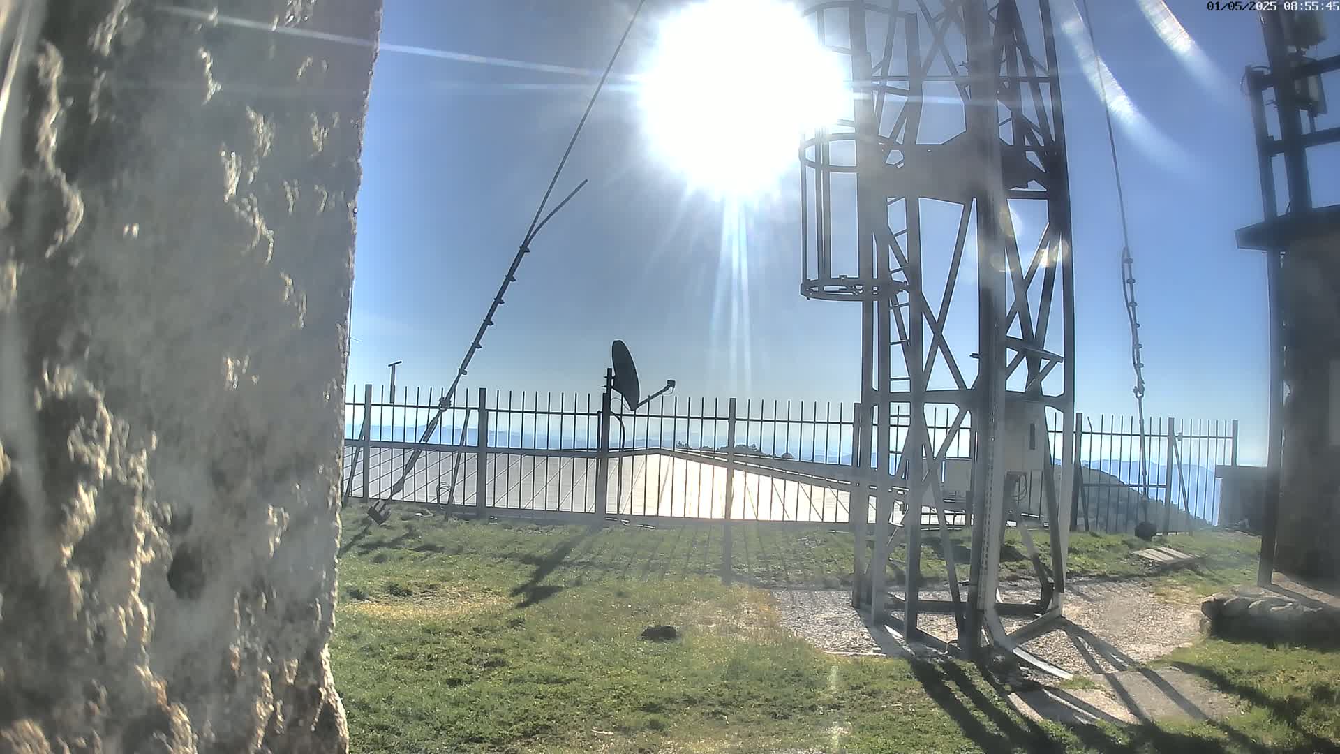 A sunny day reveals a metal tower, a satellite dish, and a metal fence overlooking a distant mountain range.