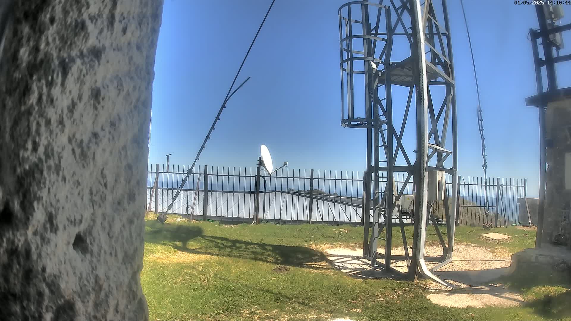 A tall metal structure with a satellite dish stands on a grassy area overlooking a fence and a distant ocean under a clear blue sky.