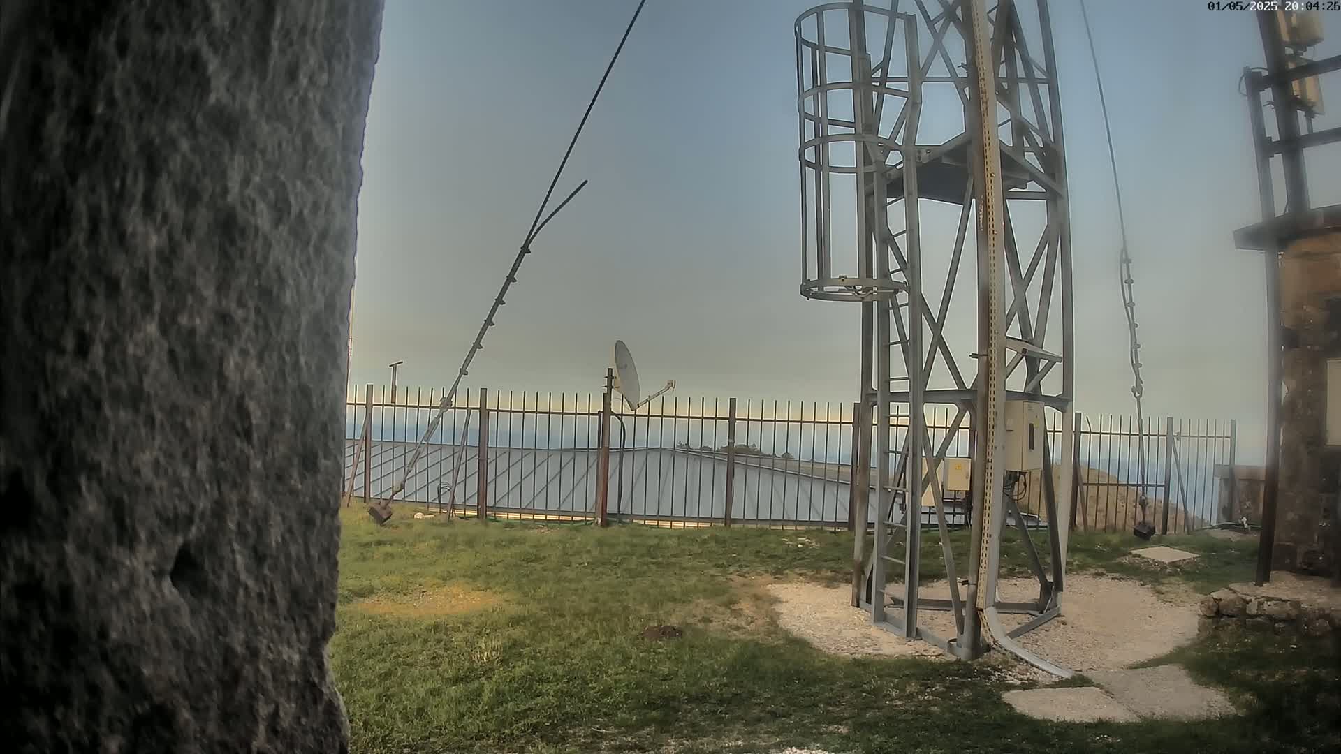 A metal communications tower with a satellite dish and antennas stands behind a metal fence overlooking a distant ocean under a cloudy sky.