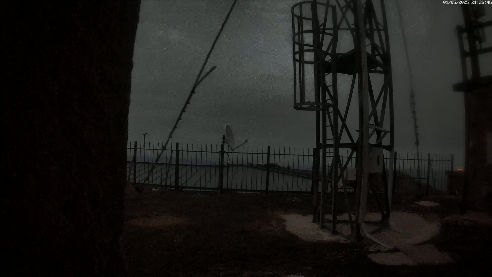 A dark, nighttime scene shows a metal tower and fence surrounding a satellite dish under a cloudy, possibly stormy sky.