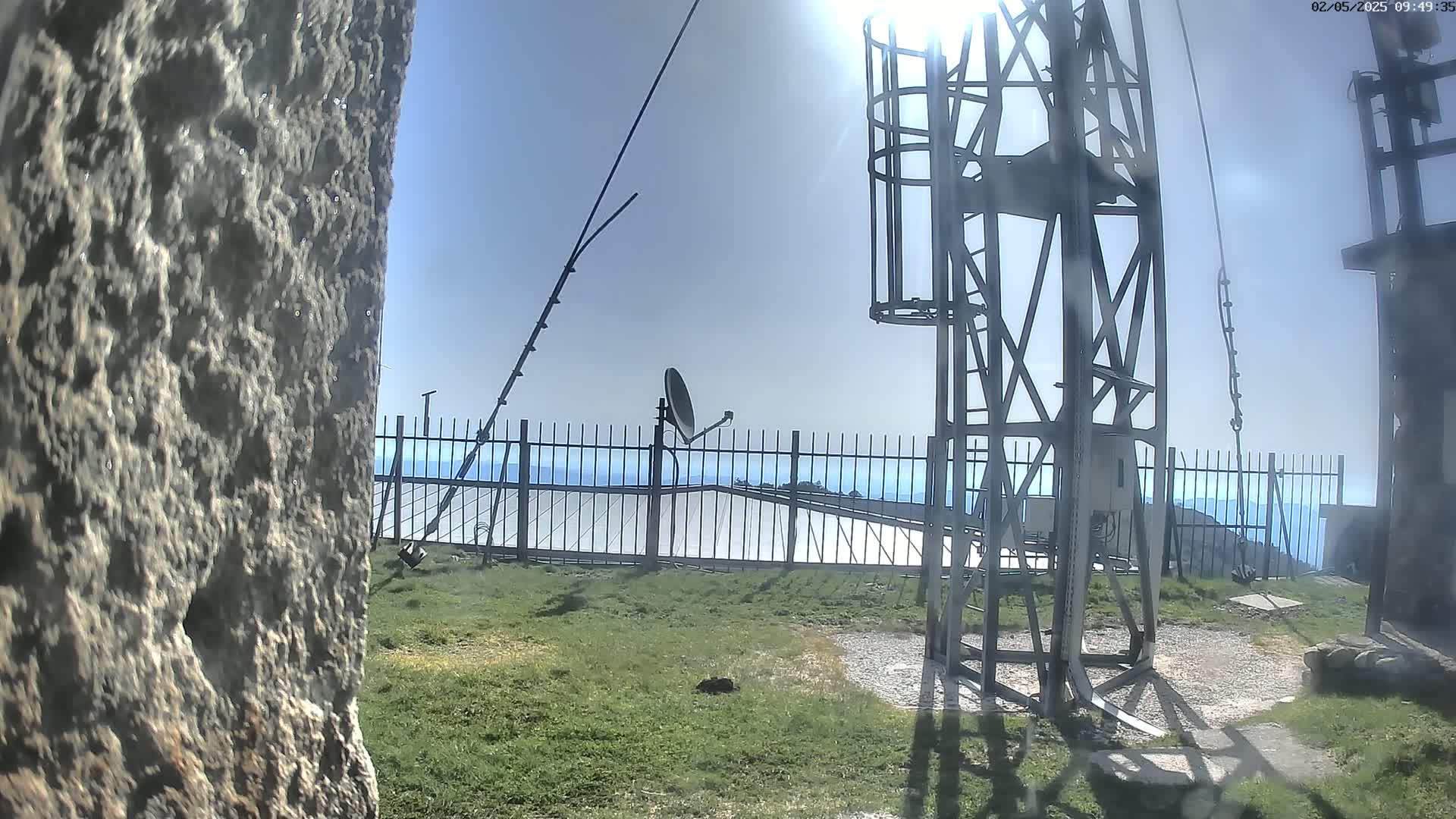 A metal communications tower and satellite dish are situated on a grassy area behind a fence overlooking a hazy, distant landscape under a clear, sunny sky.