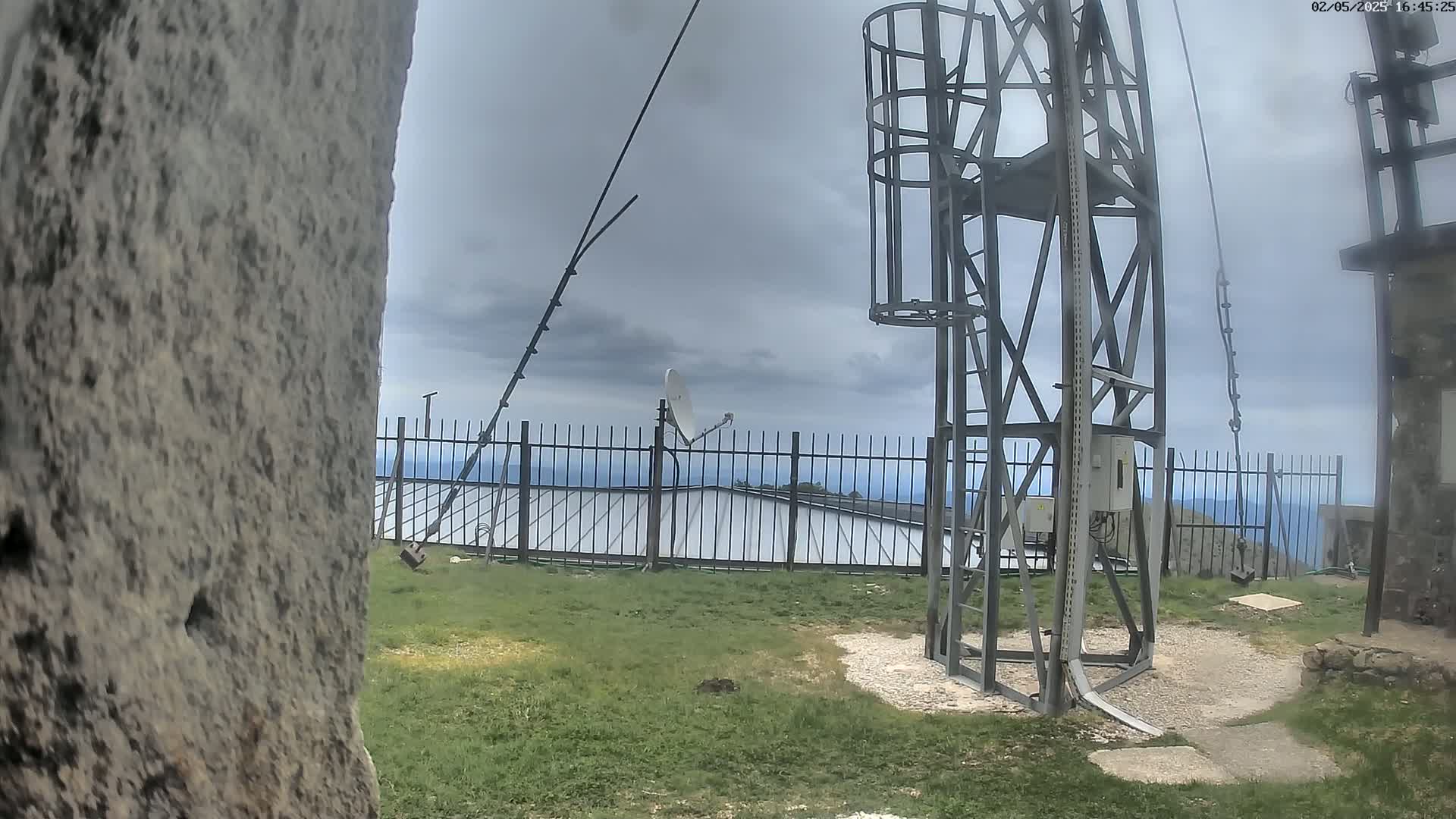 A metal tower with antennas stands on a grassy hilltop under an overcast sky, overlooking a metal fence and distant ocean.