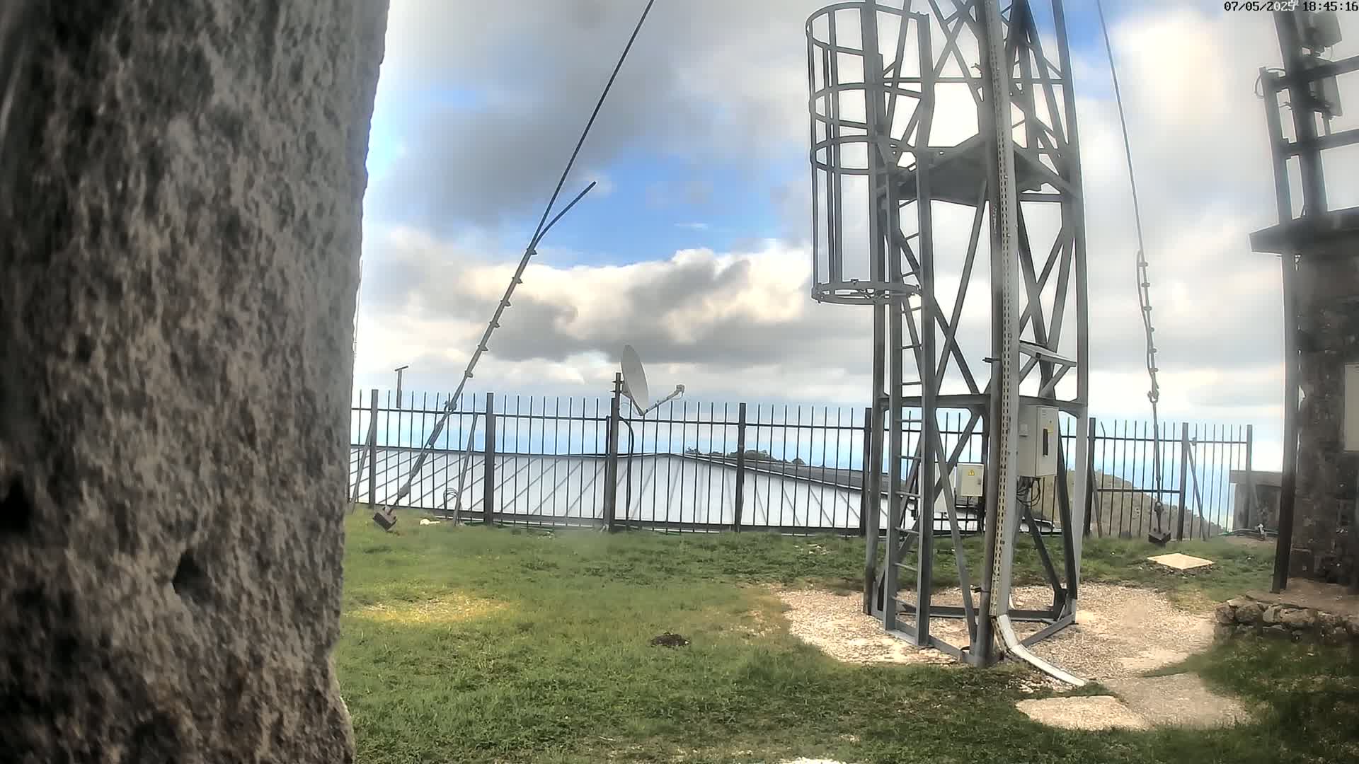 A metal tower with a cage stands on a grassy area behind a fence, under a partly cloudy sky.