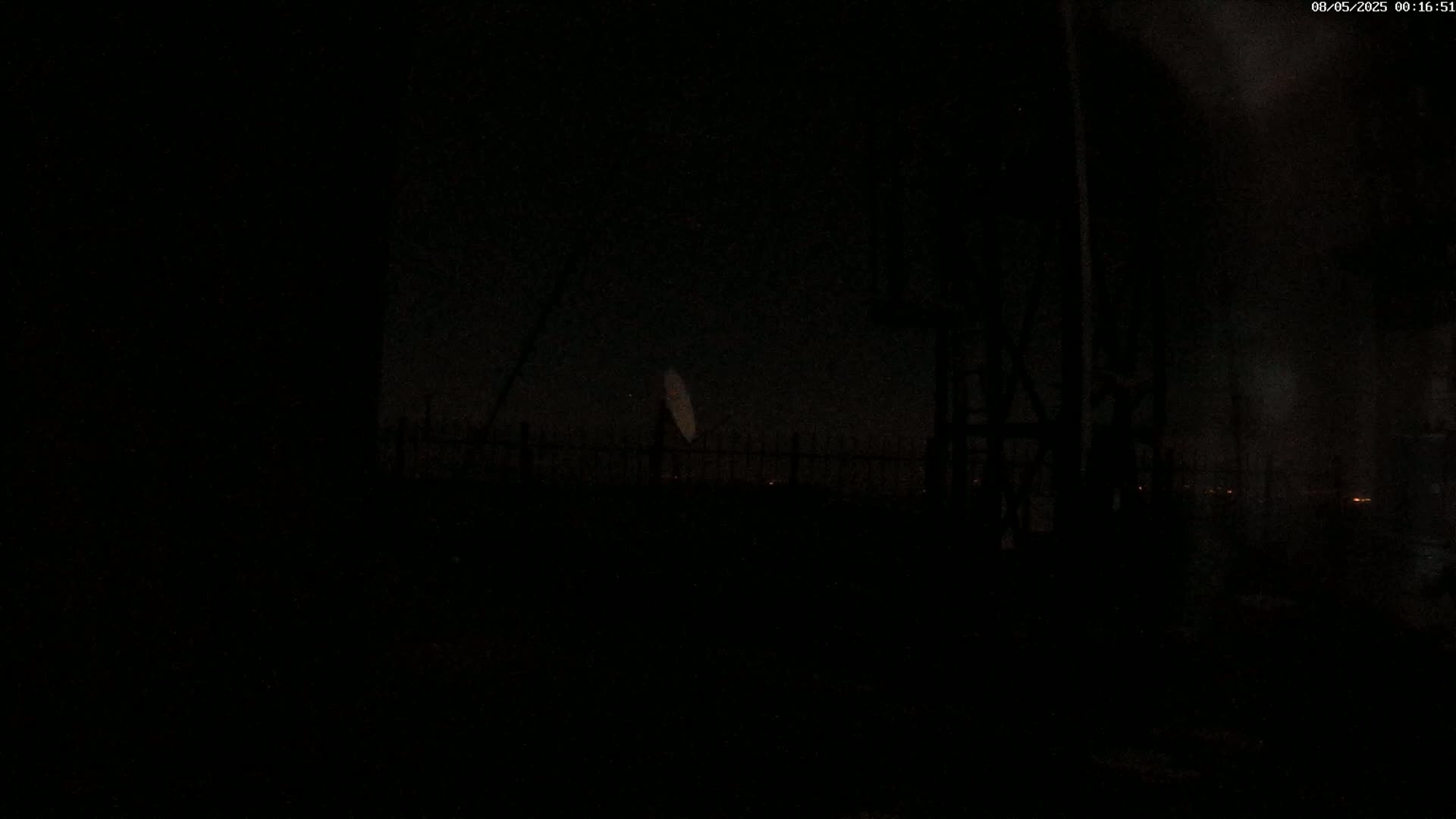 A dark, nighttime scene shows a large satellite dish behind a fence, with dimly lit areas visible in the distance.