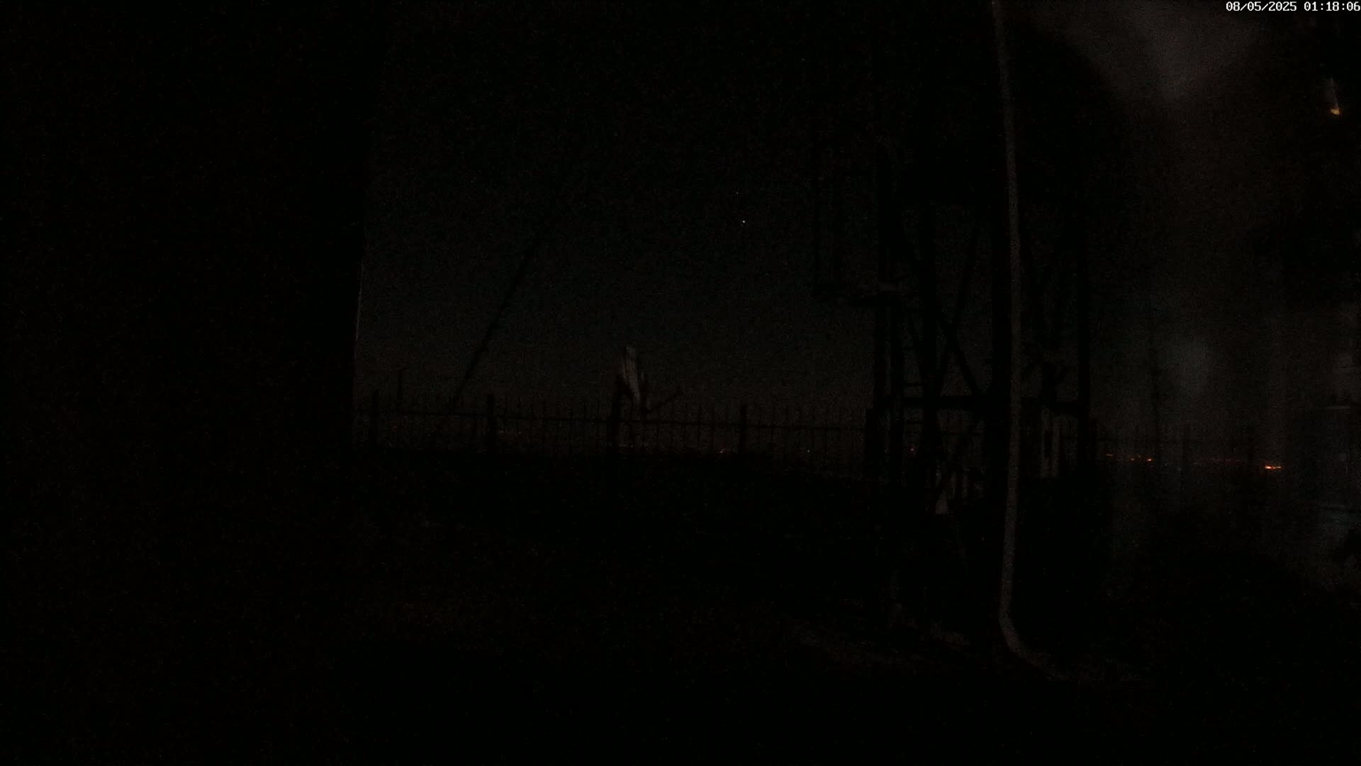 A dark, nighttime scene shows a dimly lit fence and structure, with a few distant lights visible in the background under a mostly clear sky.