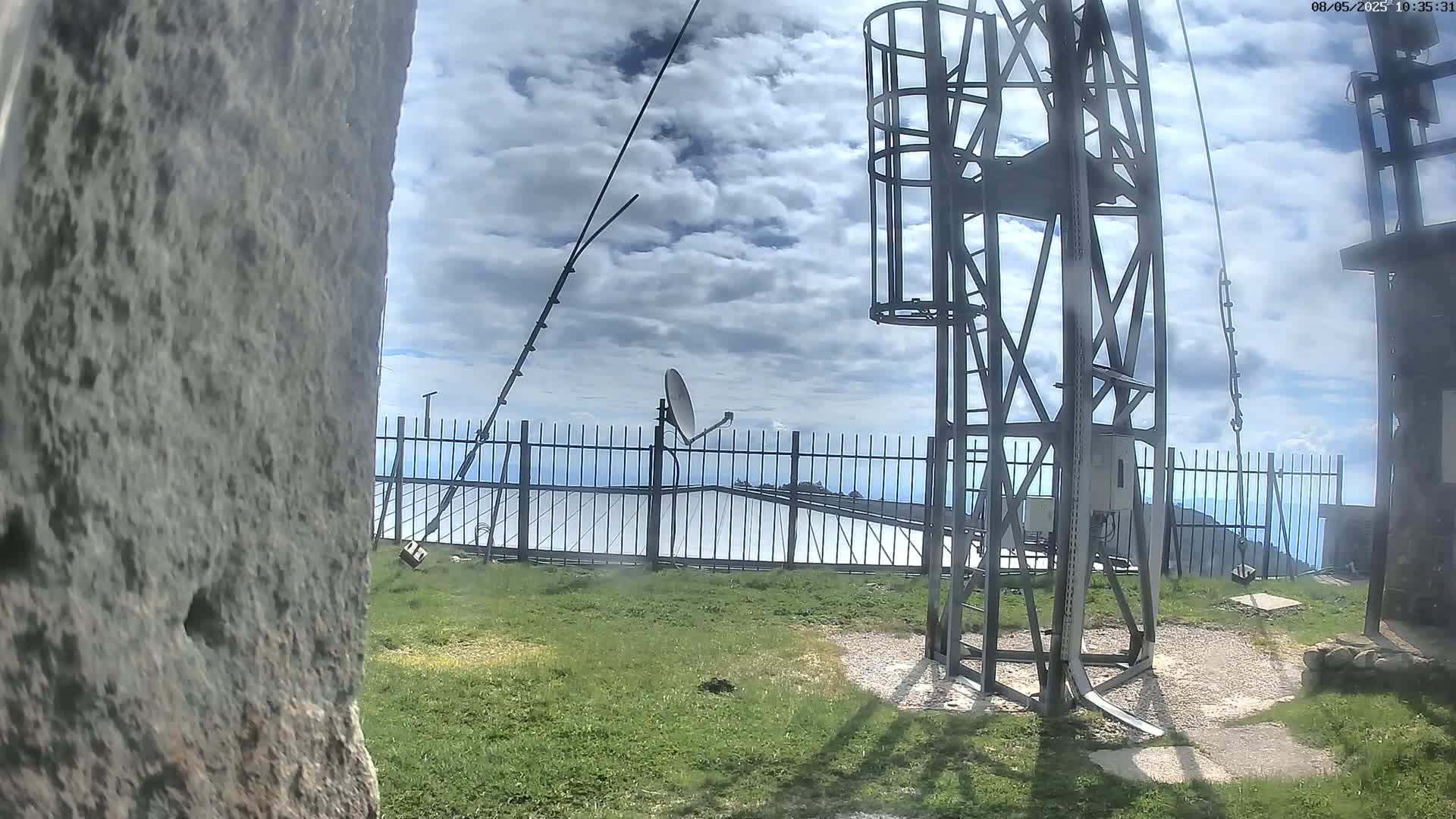 A metal communications tower with a satellite dish and antenna stands on a grassy area behind a fence, under a partly cloudy sky.
