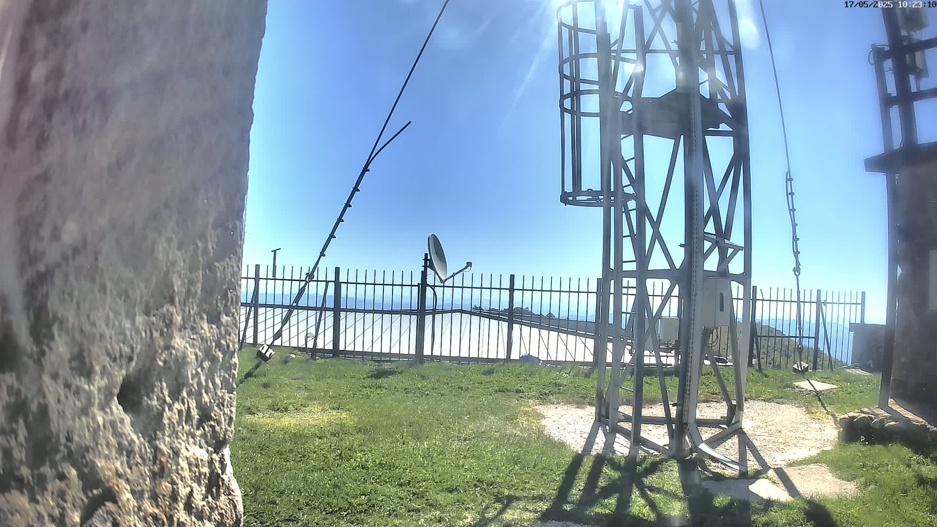 A metal communications tower stands on a grassy area overlooking a hazy ocean under a clear blue sky.