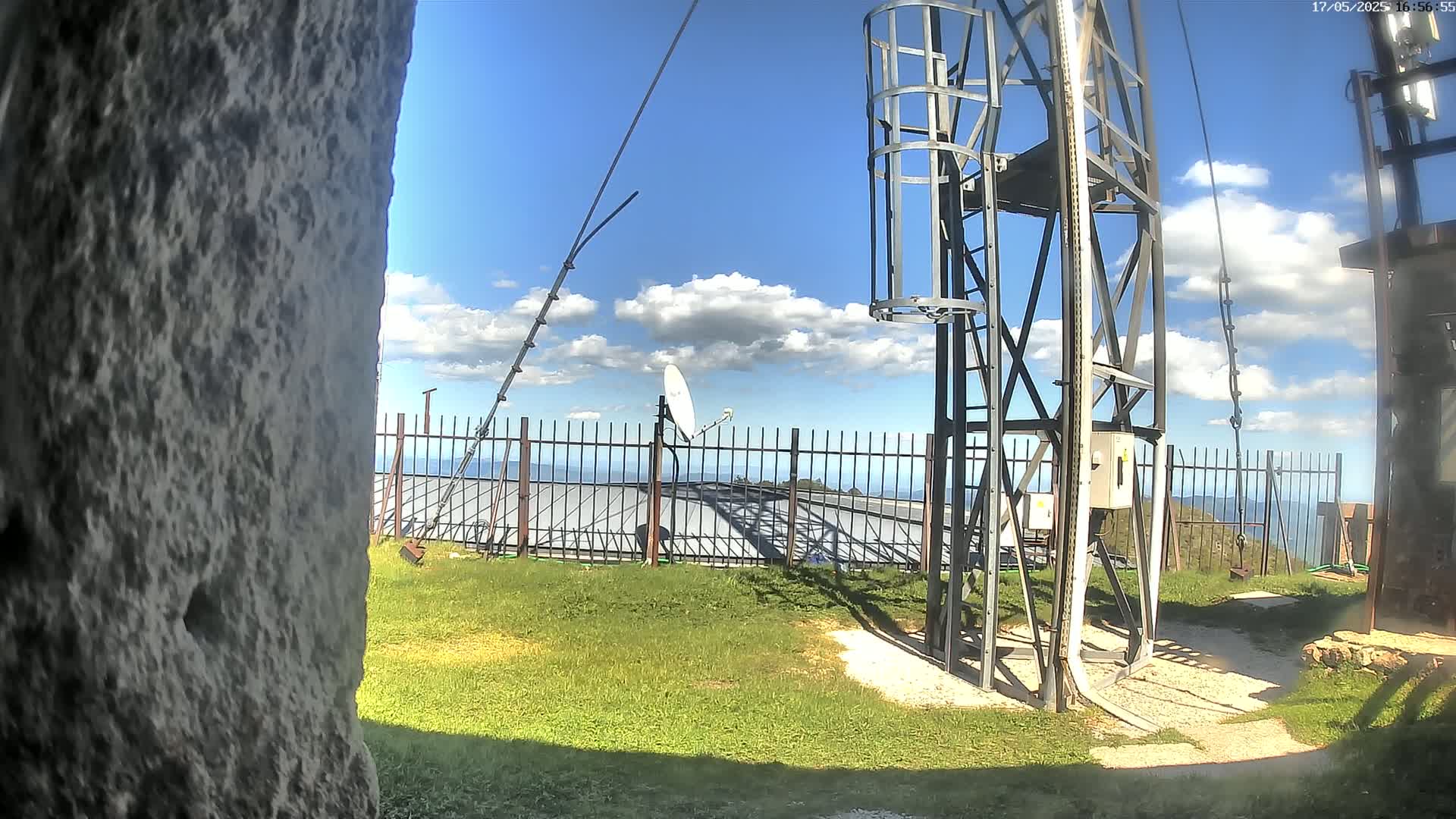 A metal communications tower with a satellite dish stands on a grassy area with a metal fence overlooking a distant ocean under a partly cloudy blue sky.