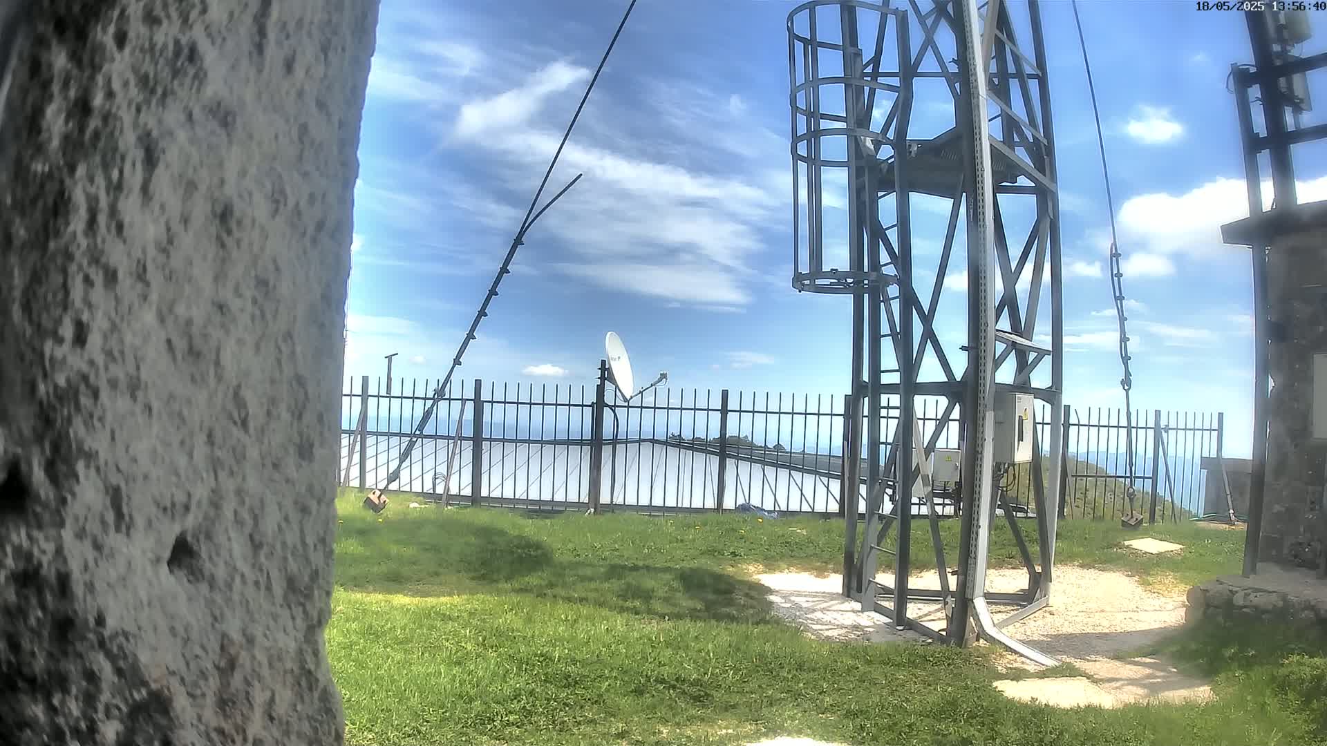 A metal tower with a satellite dish and solar panels is situated behind a fence on a grassy area under a partly cloudy sky.