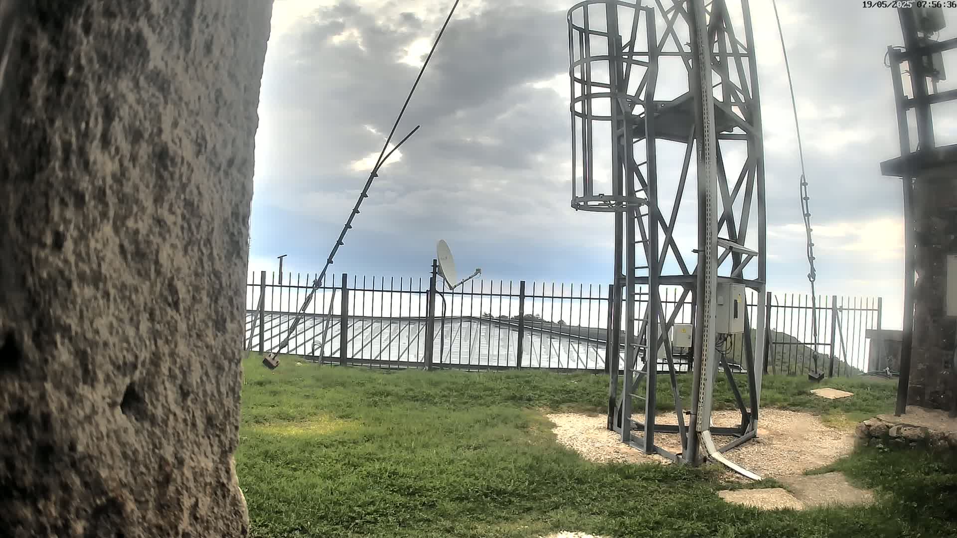 A metal tower with a satellite dish and a fence stands on a grassy area under a cloudy sky.