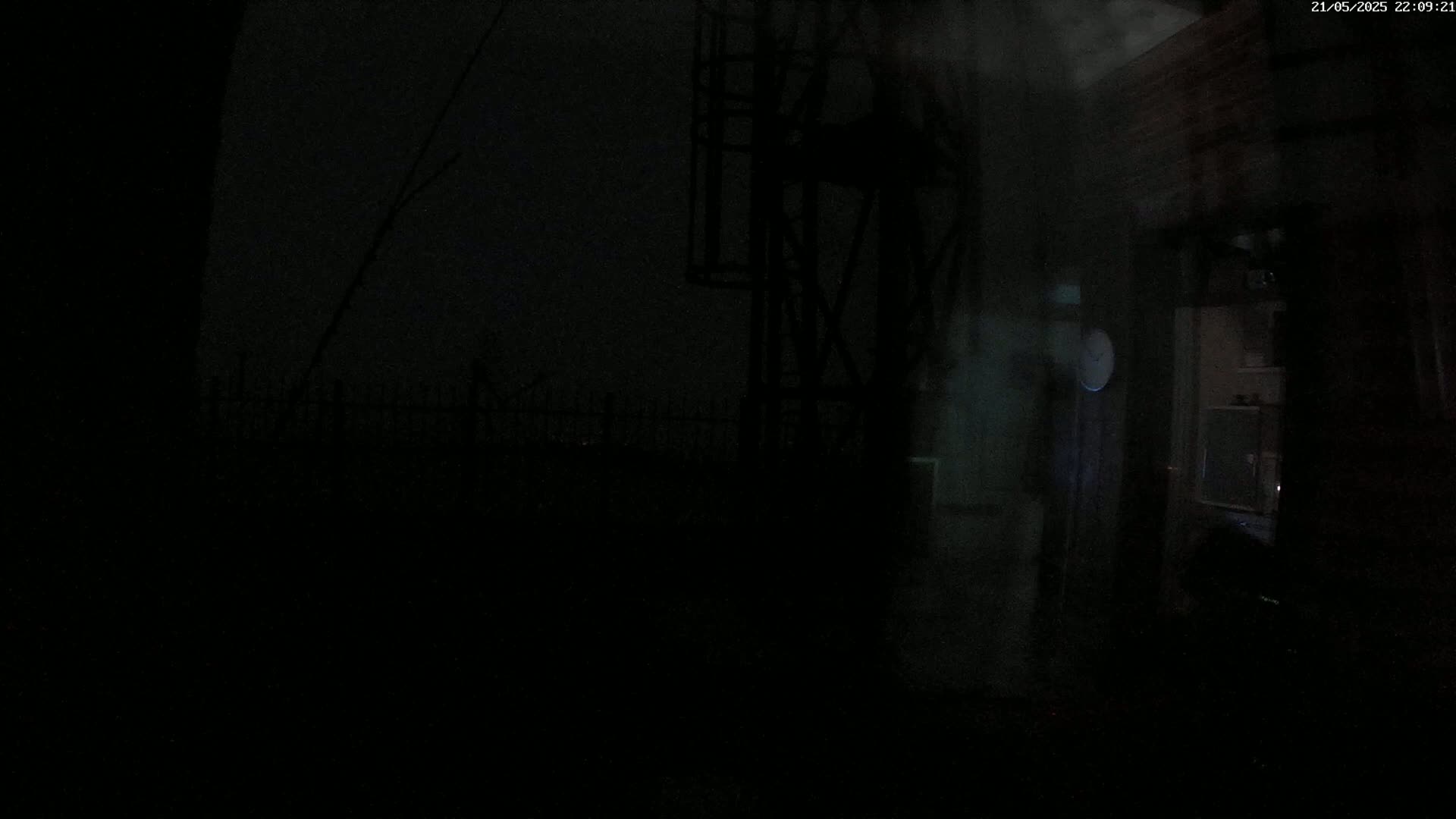 A dark, nighttime scene shows a metal structure next to a building, with a fence visible in the distance under a hazy sky.