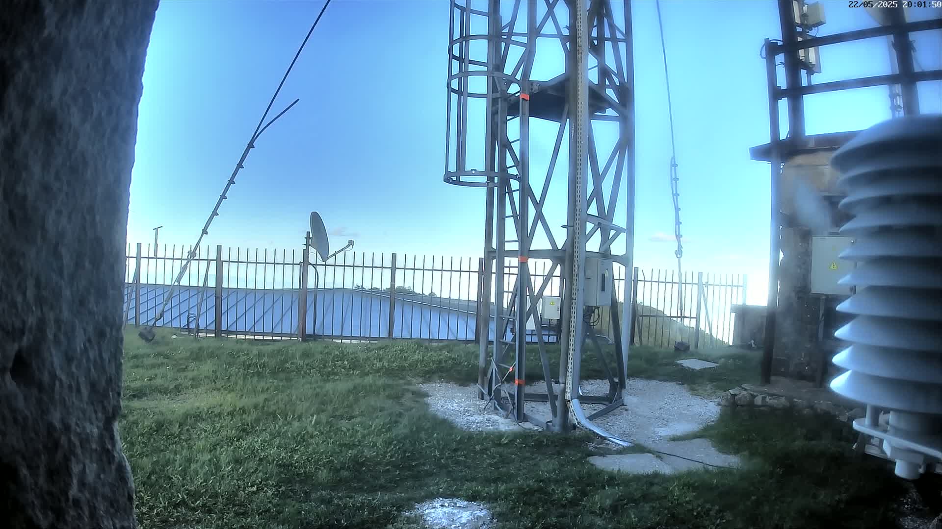 A metal tower with a cage-like structure stands on grassy ground under a clear blue sky, near a fence, a satellite dish, and a weather station.