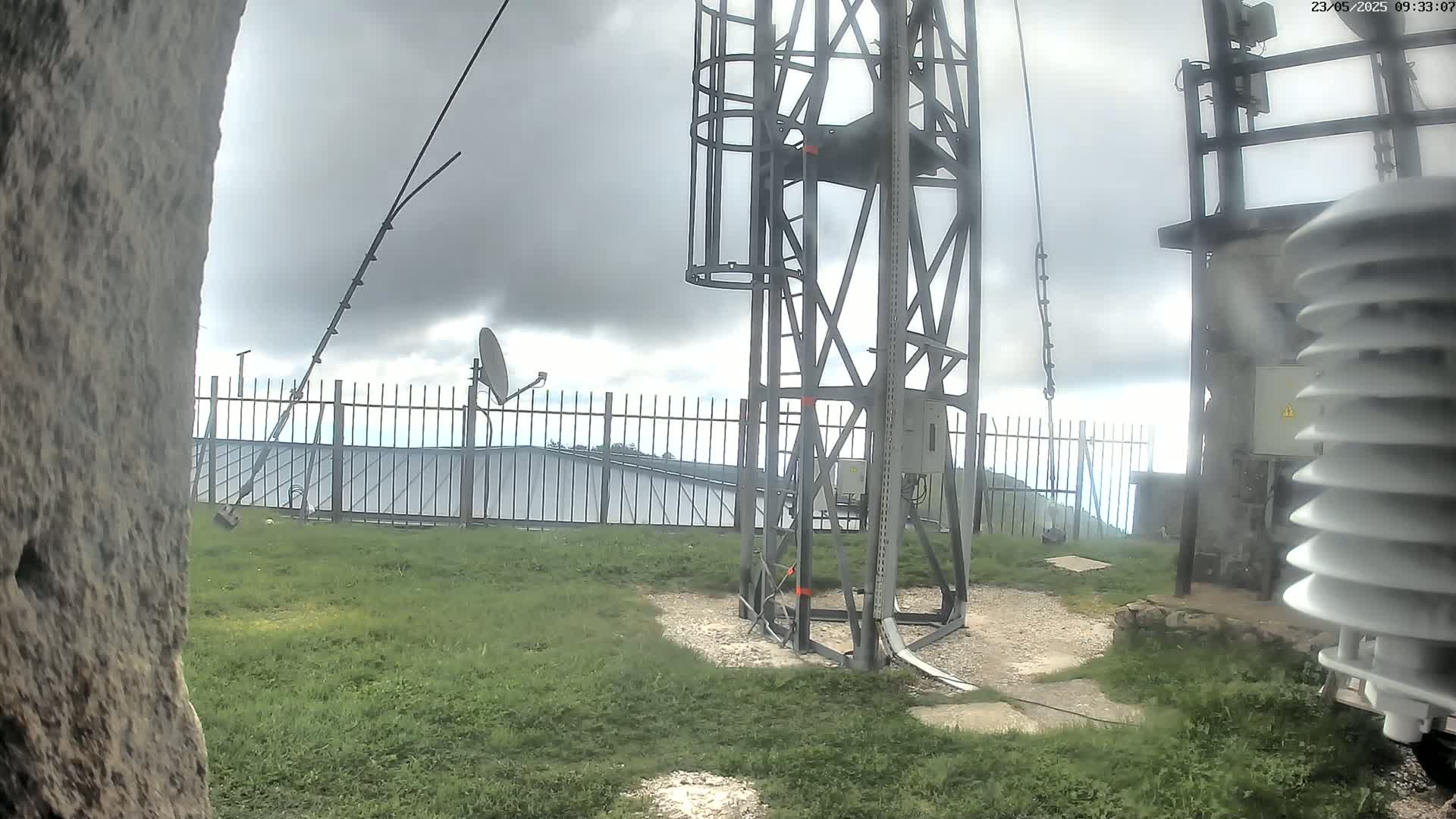 A metal tower with a cage-like structure stands on a grassy area under a cloudy sky, near a fence, a satellite dish, and other equipment.