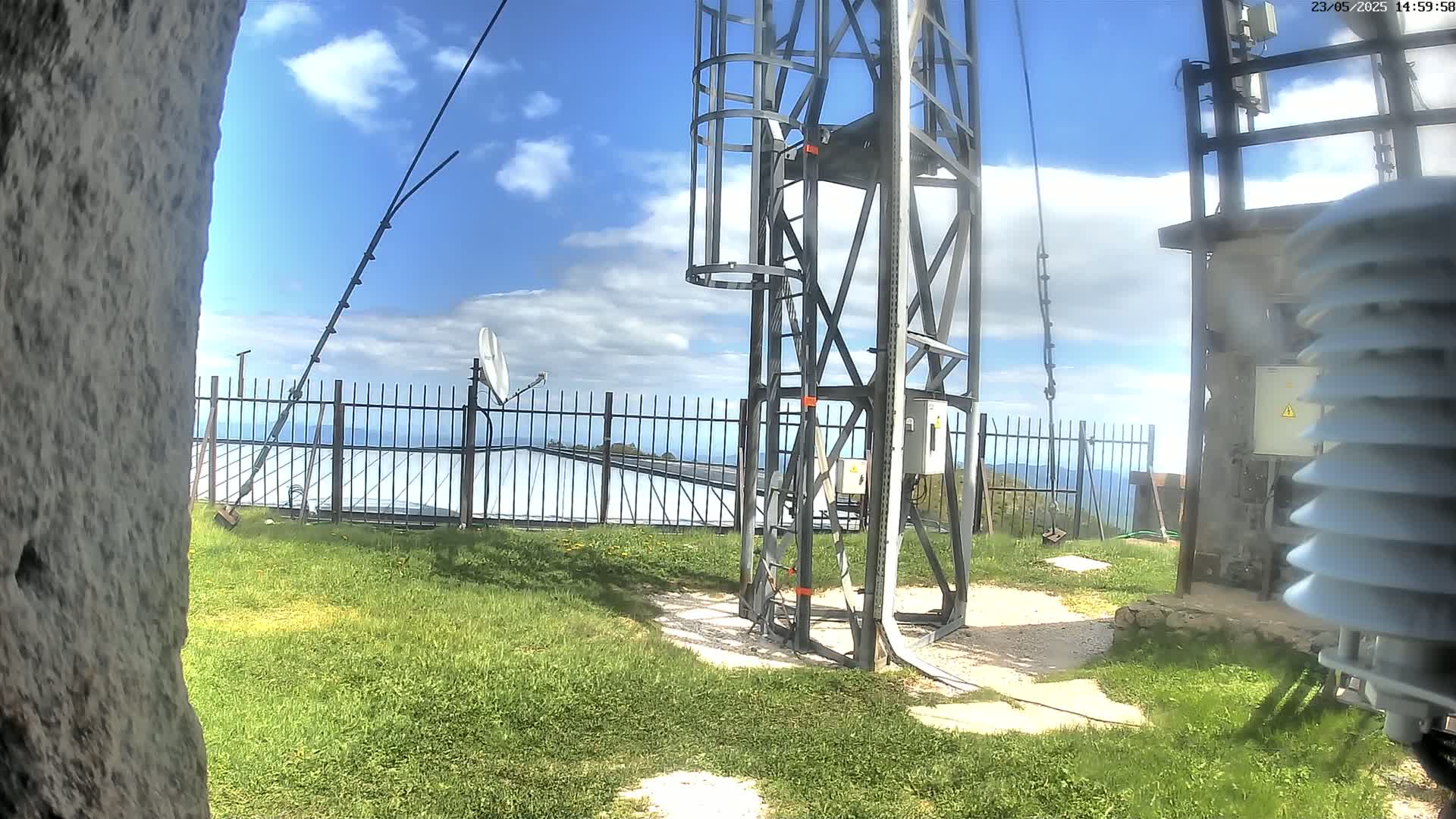 A metal tower with a ladder and various antennas stands on grassy land behind a metal fence, under a partly cloudy sky.